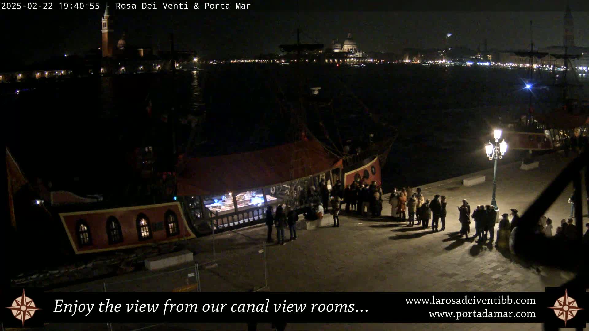 A nighttime view of a canal, with a docked ship and a crowd of people standing nearby, under a dark sky.
