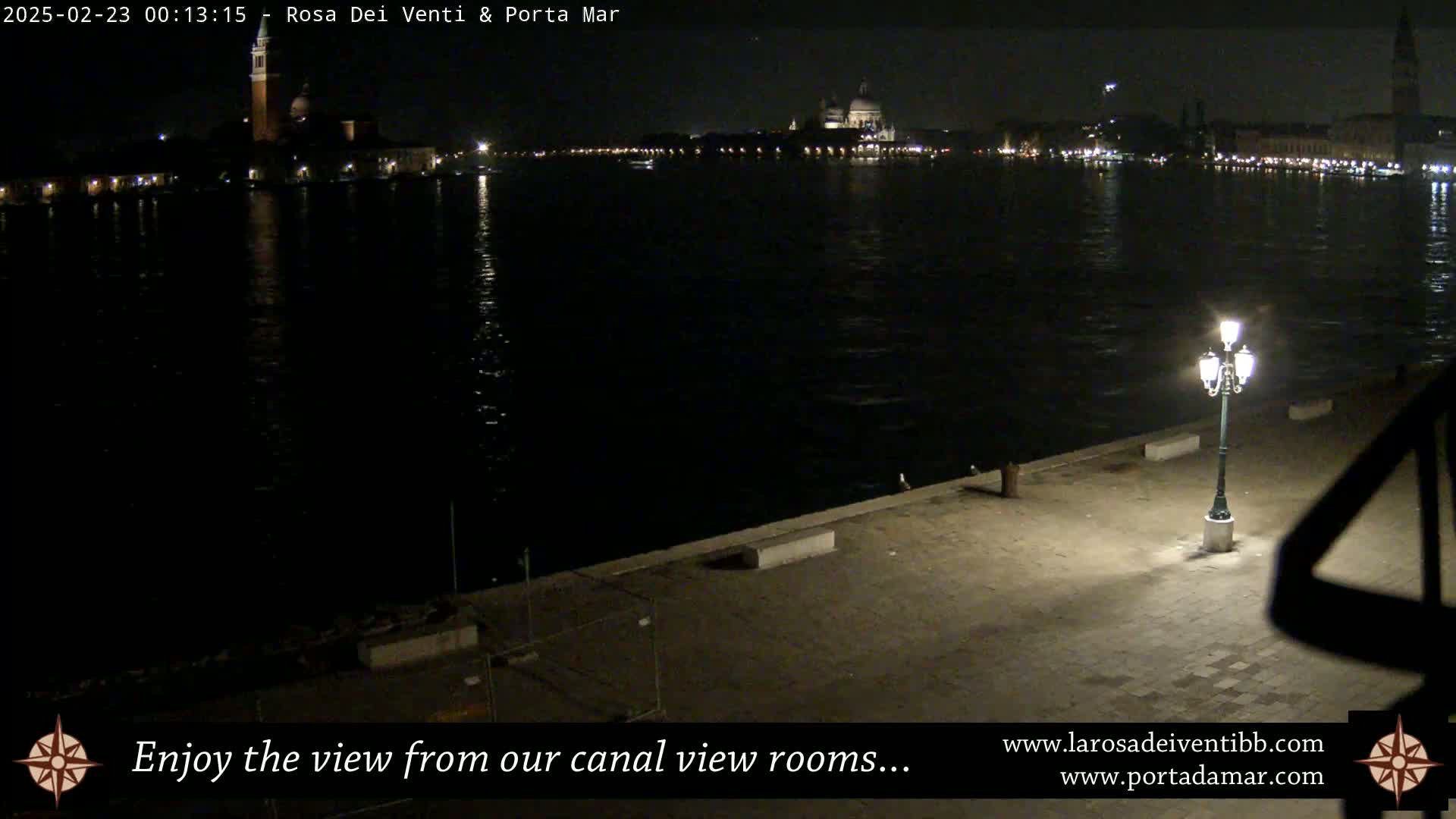 A nighttime view of a calm canal shows a cityscape with illuminated buildings across the water, and a single lamppost on a paved waterfront area.