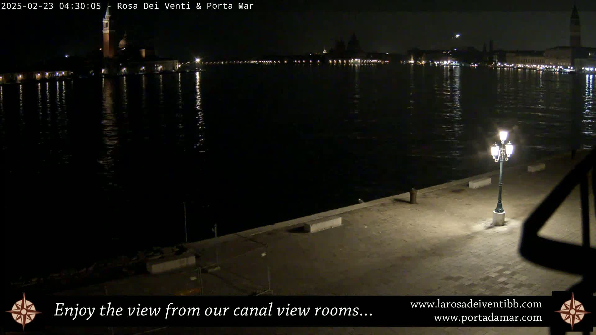 A nighttime view across a calm canal shows a city skyline with lights reflected in the water, and a single lit lamppost on a stone embankment in the foreground.