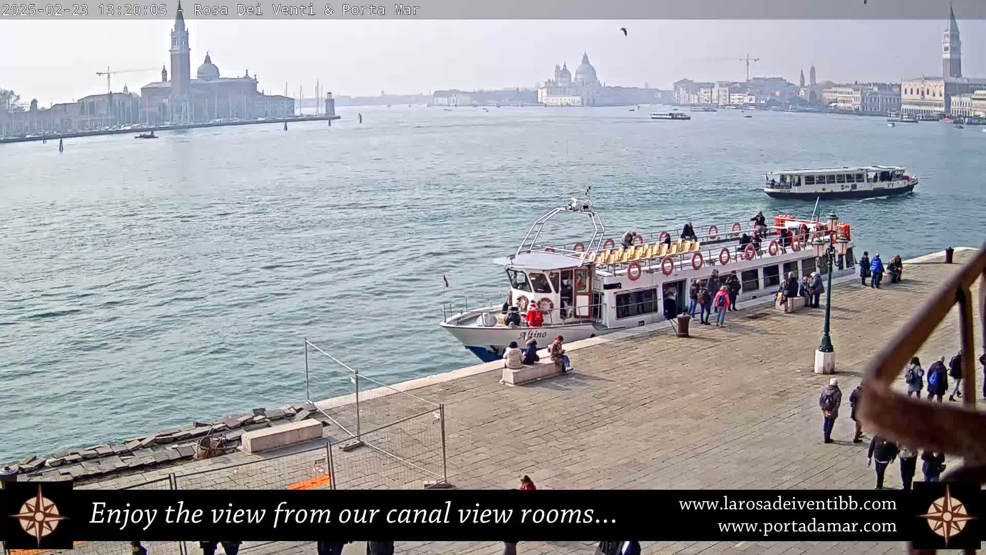 A canal-side view in Venice on a hazy day shows a docked passenger boat, people walking on a paved area, and distant buildings across the water.