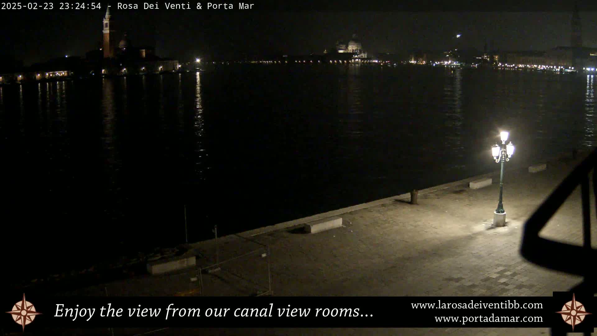 A nighttime view across a dark canal shows illuminated buildings on the far shore, a single lit lamppost in the foreground, and a paved walkway.