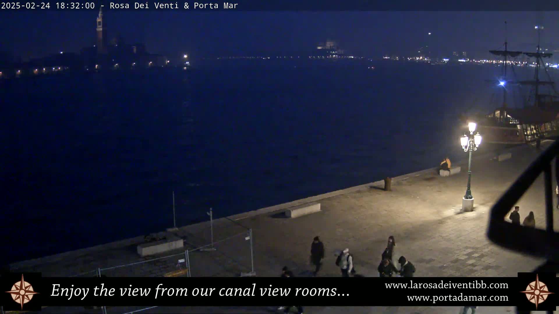 A nighttime view of a canal, showing a cityscape across the water, a docked ship, and several people walking along a paved waterfront.