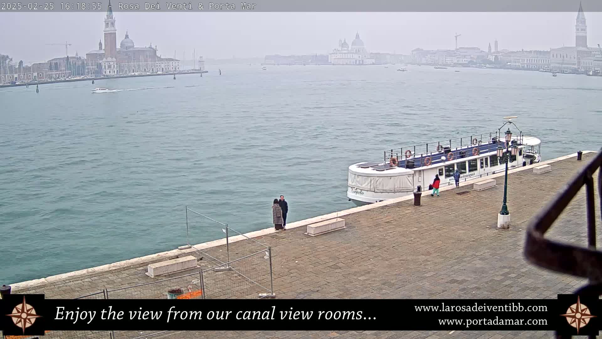A hazy view of a canal with a docked passenger boat, a couple standing nearby, and a distant cityscape in the background.