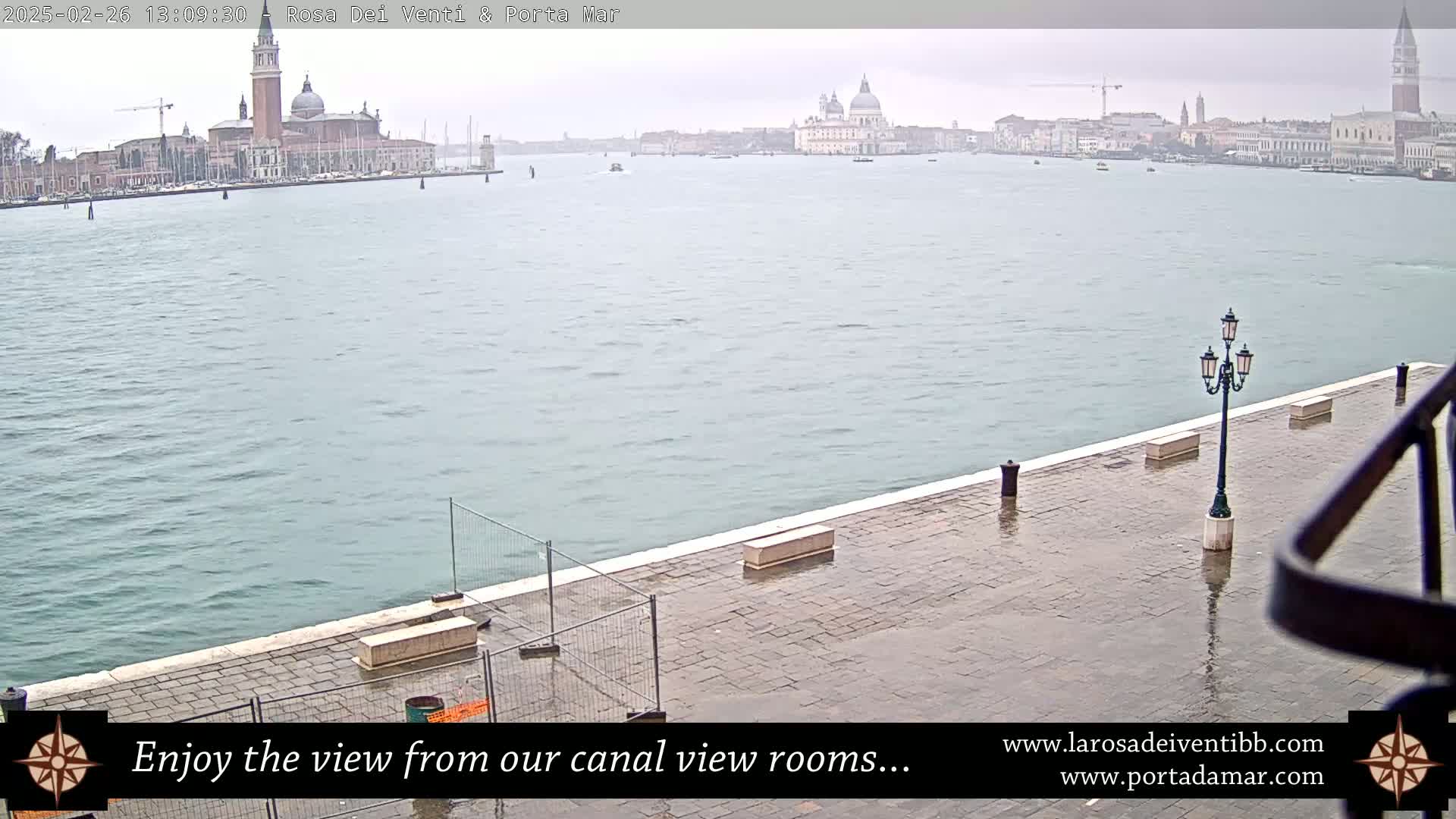 A wet, paved waterfront area overlooks a canal with a distant view of buildings and a church under an overcast sky.