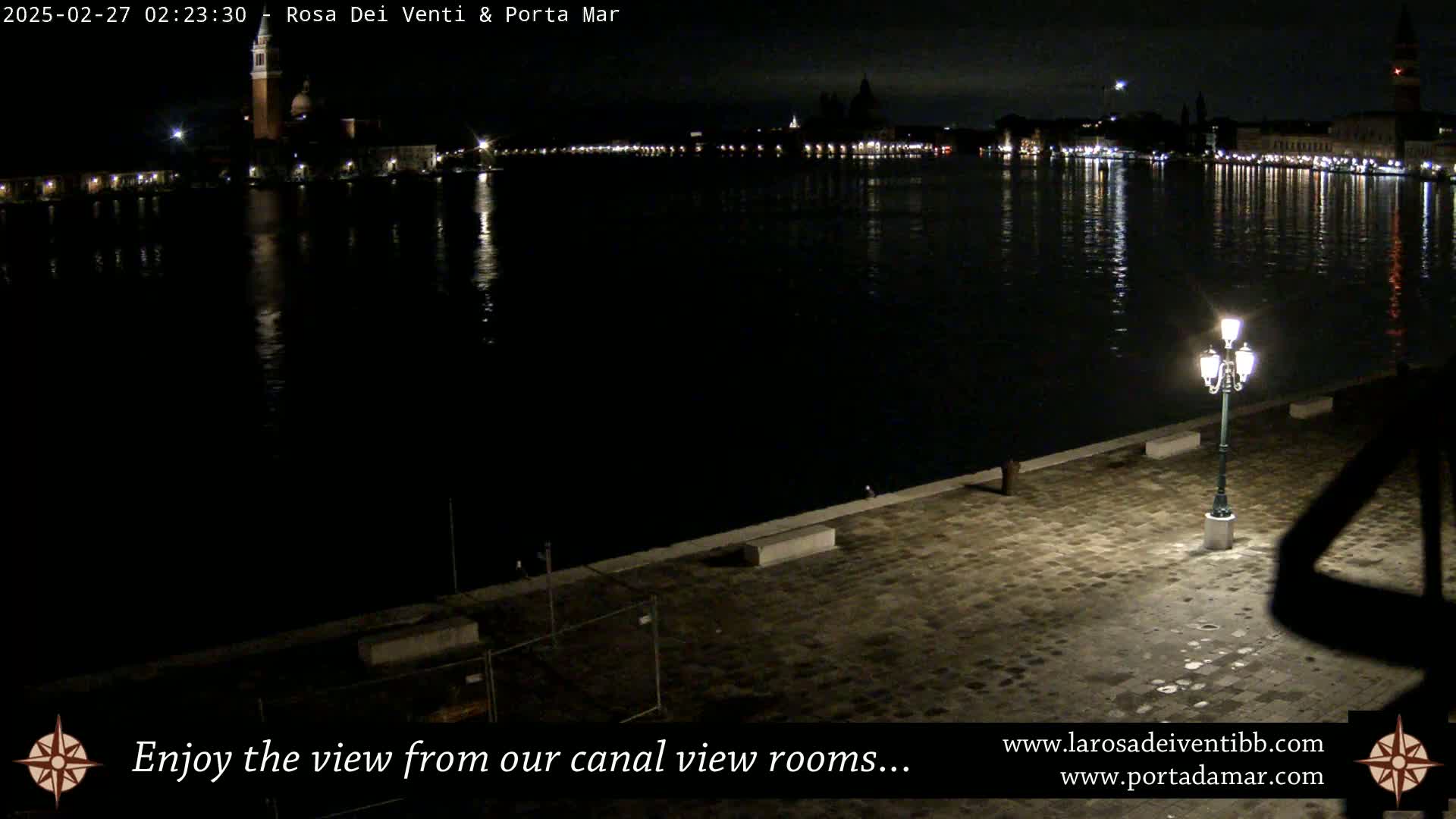 A nighttime view of a calm canal reflecting lights from buildings across the water, with a single lamppost illuminating a paved area in the foreground.