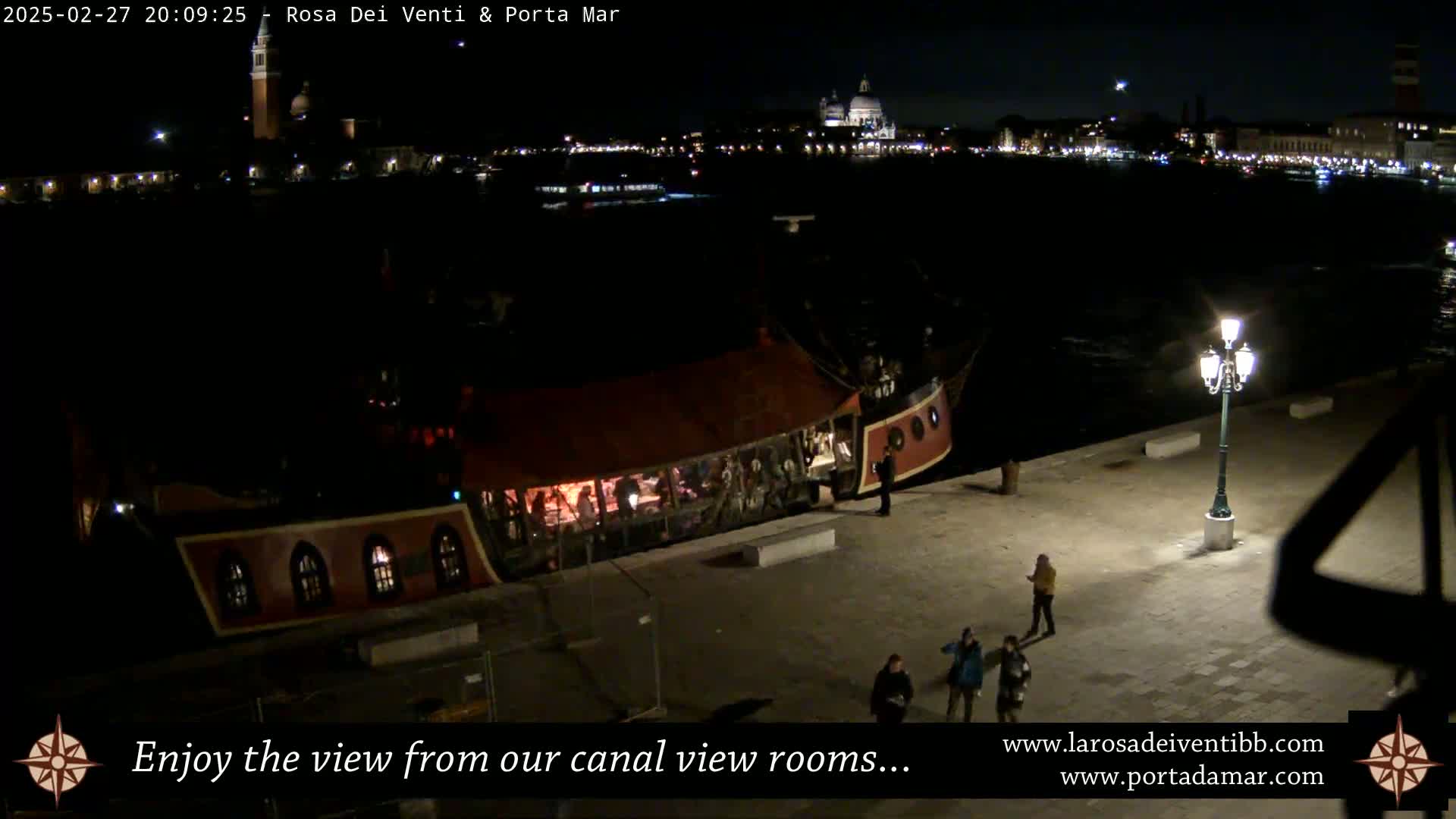 A pirate ship-like vessel is docked at night near a waterfront, with city lights and buildings visible across the water under a clear sky.
