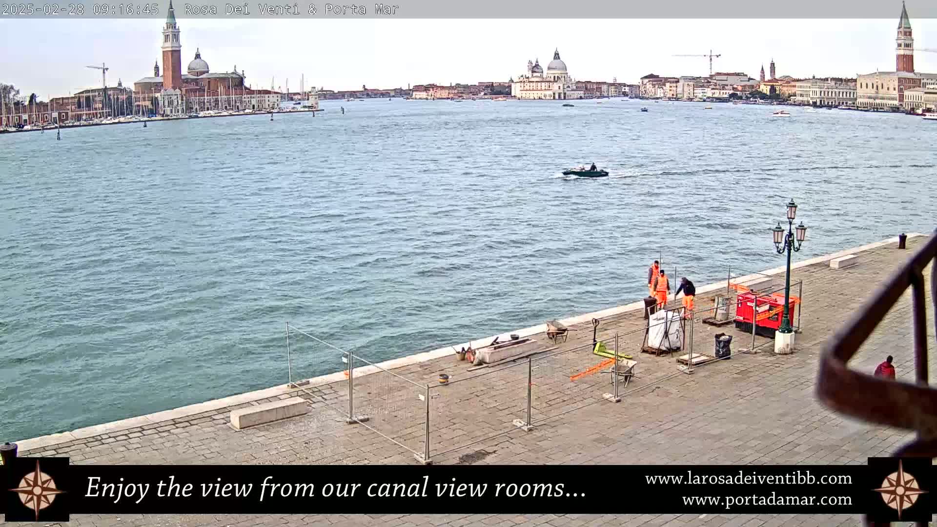 A waterfront view of Venice, Italy, on an overcast day shows workers on a paved area near a calm canal with boats, and a distant cityscape including churches.