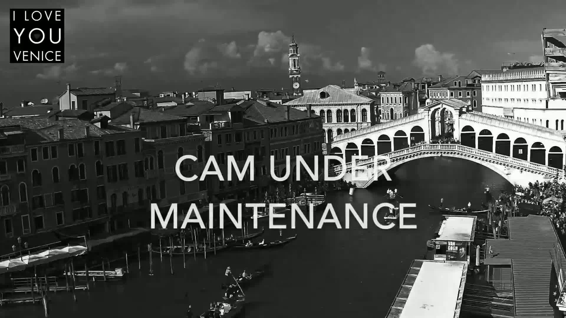 A black and white, high-angle view shows a Venetian canal with gondolas, a bridge, and buildings under a partly cloudy sky.