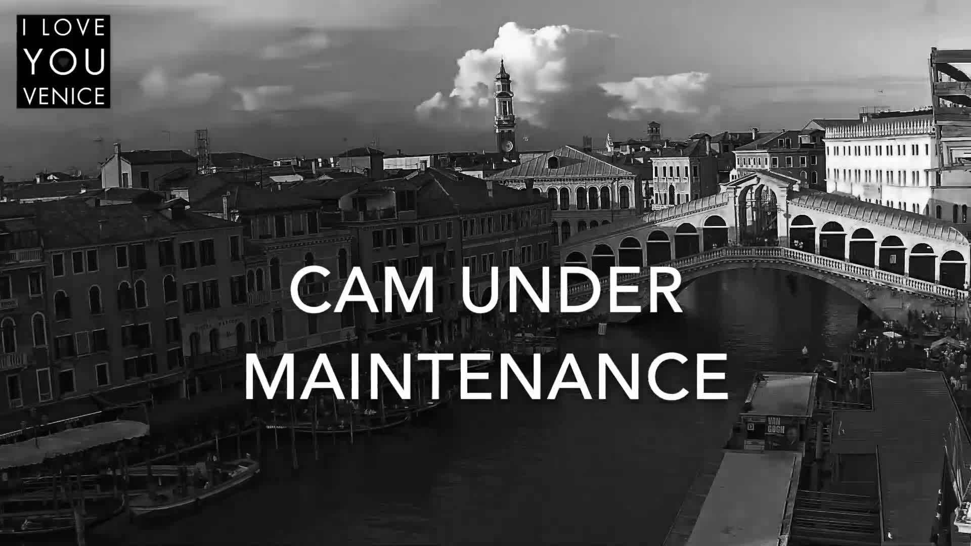 A black and white aerial view of a canal in Venice, Italy, under a partly cloudy sky, shows a bridge spanning the waterway with buildings lining the canal's edges.