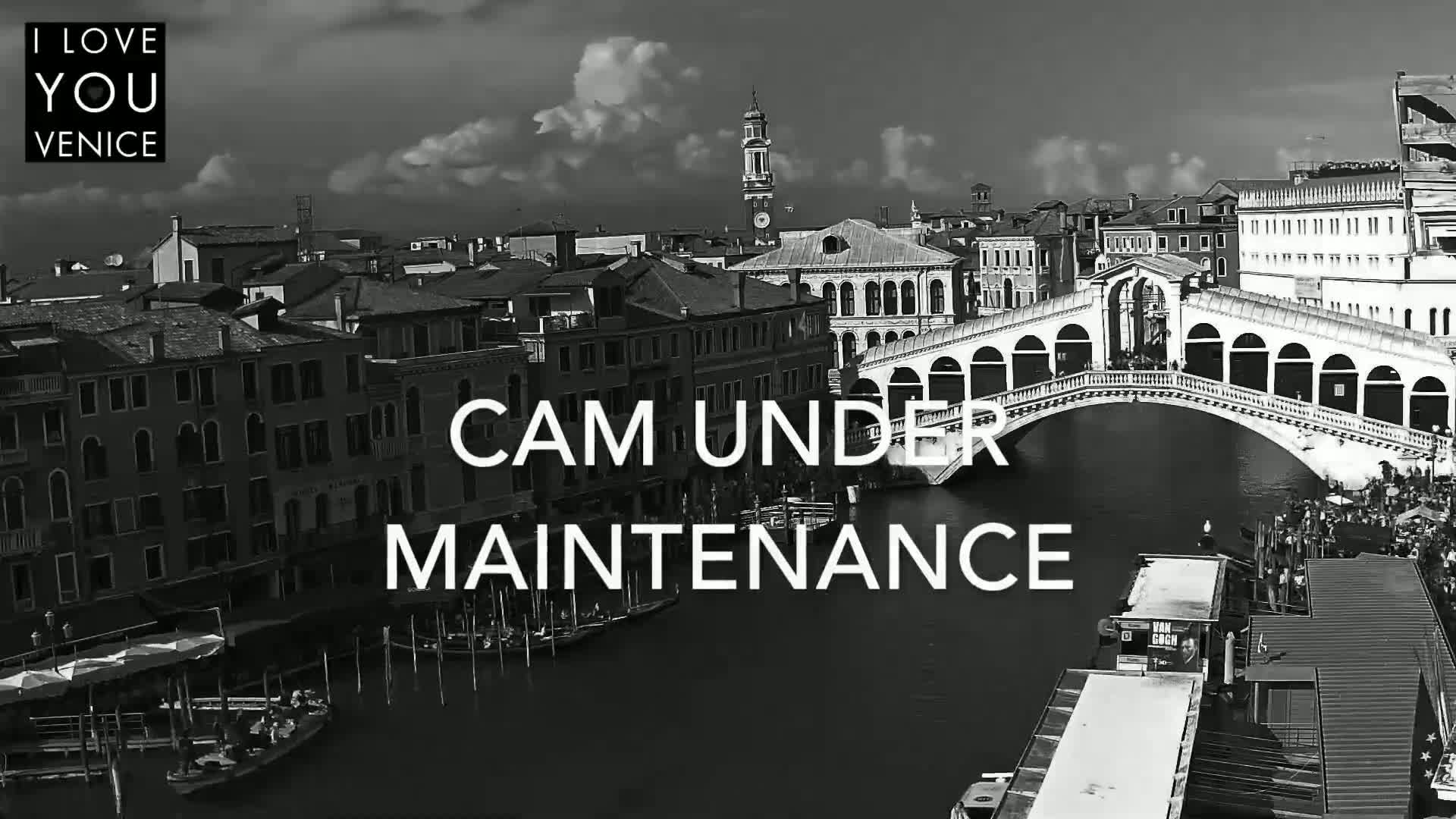 A black and white, high-angle view shows a canal in Venice, Italy, with a bridge and many buildings, under a partly cloudy sky.