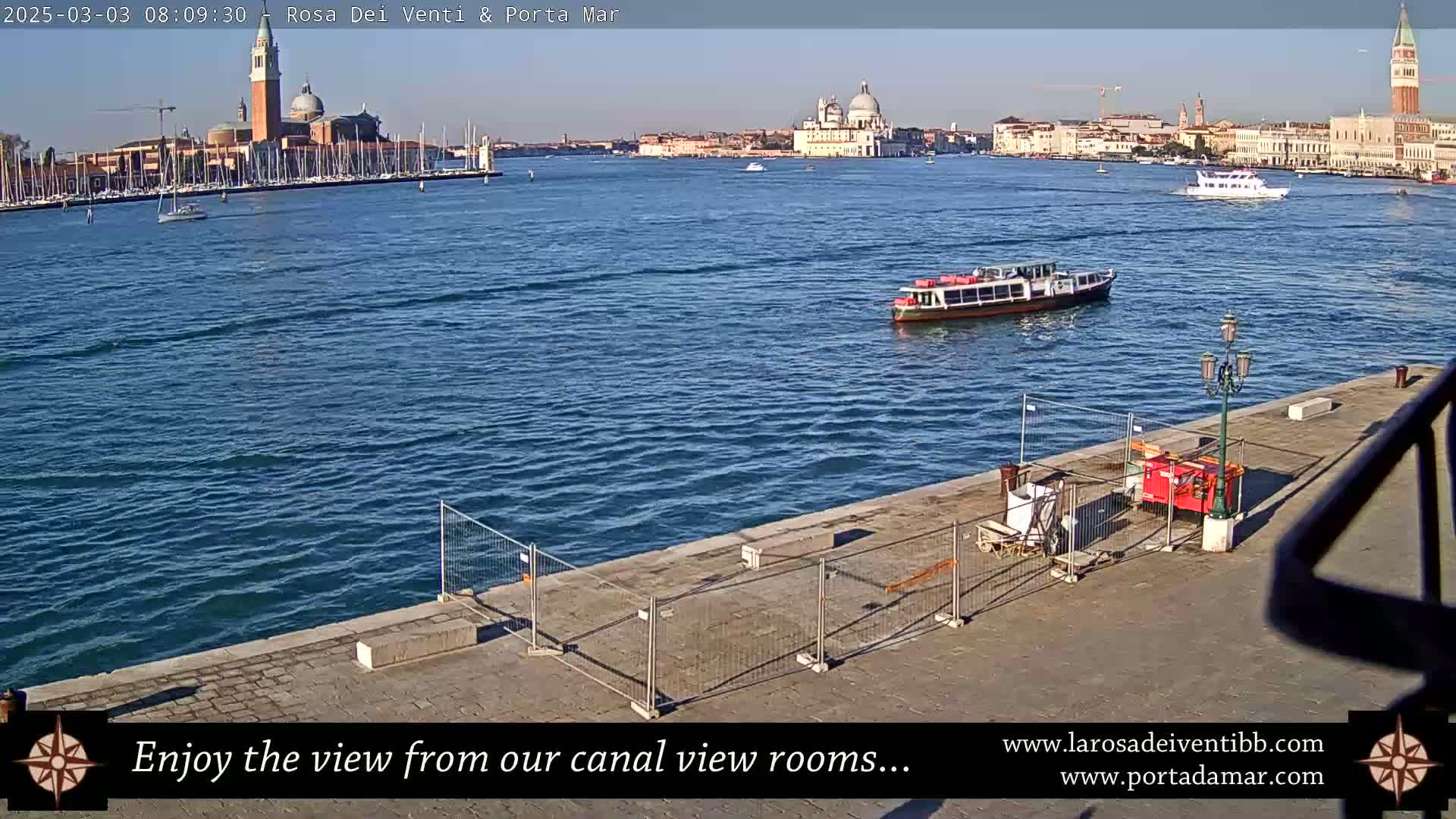 A waterfront view of Venice, Italy on a sunny day, shows a calm canal with boats, and a cityscape including a church with a dome and bell tower in the background, with construction fencing in the foreground.