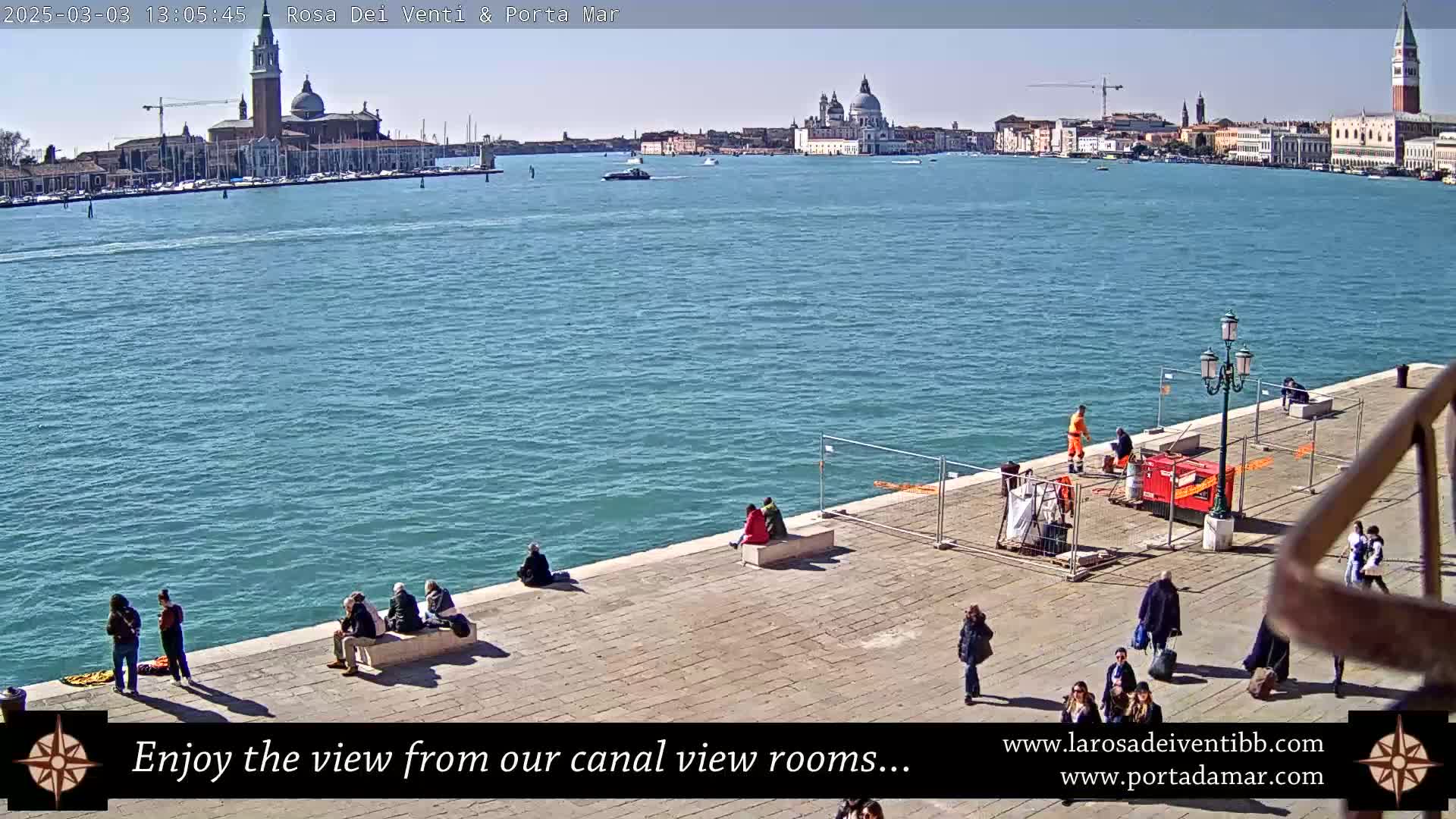 A sunny day in Venice shows a canal view with several people sitting and walking along a stone embankment, overlooking a wide expanse of water with buildings and boats in the distance.