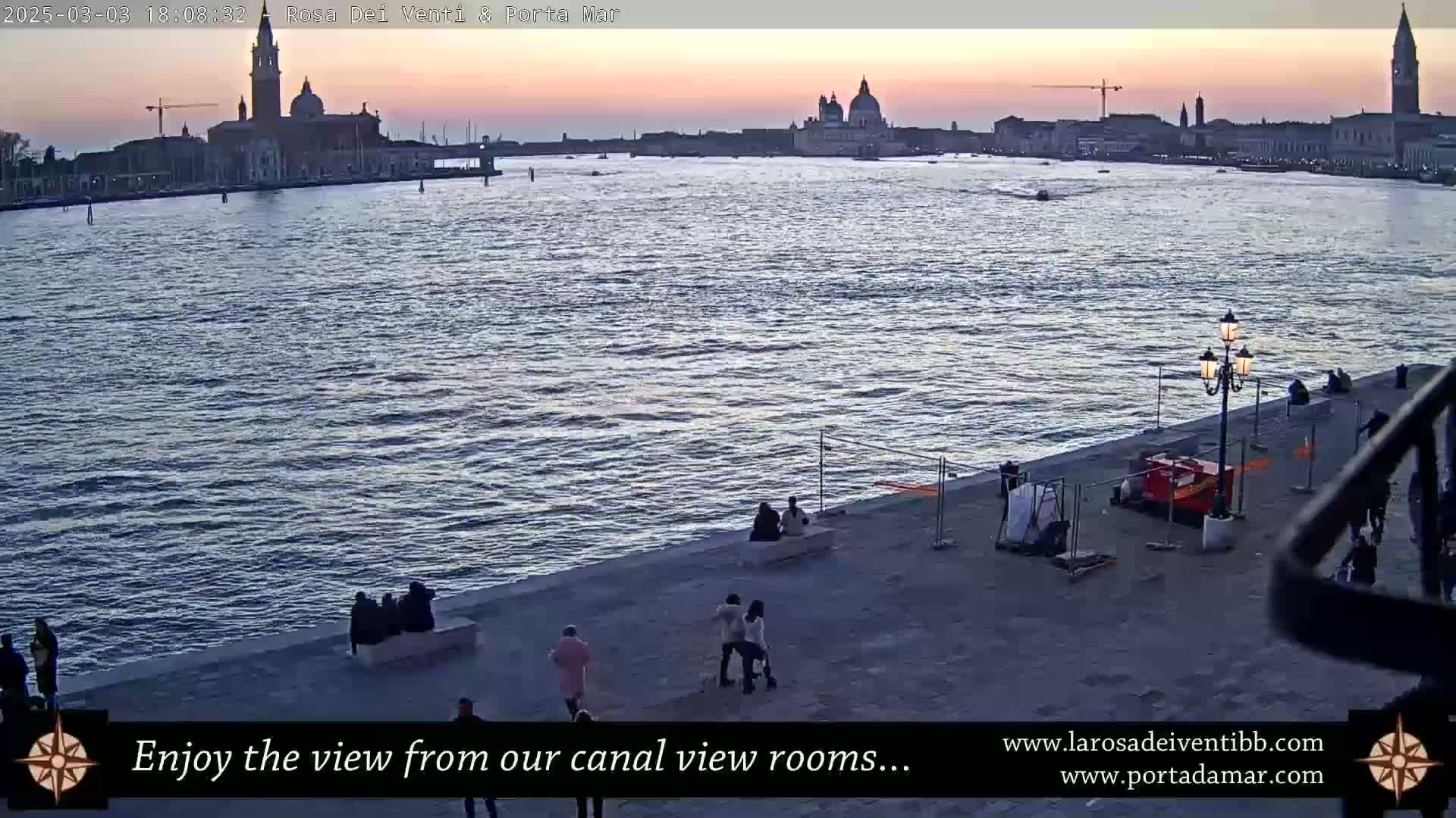 A wide shot of a calm body of water at sunset, with a city skyline and several people along a waterfront promenade.
