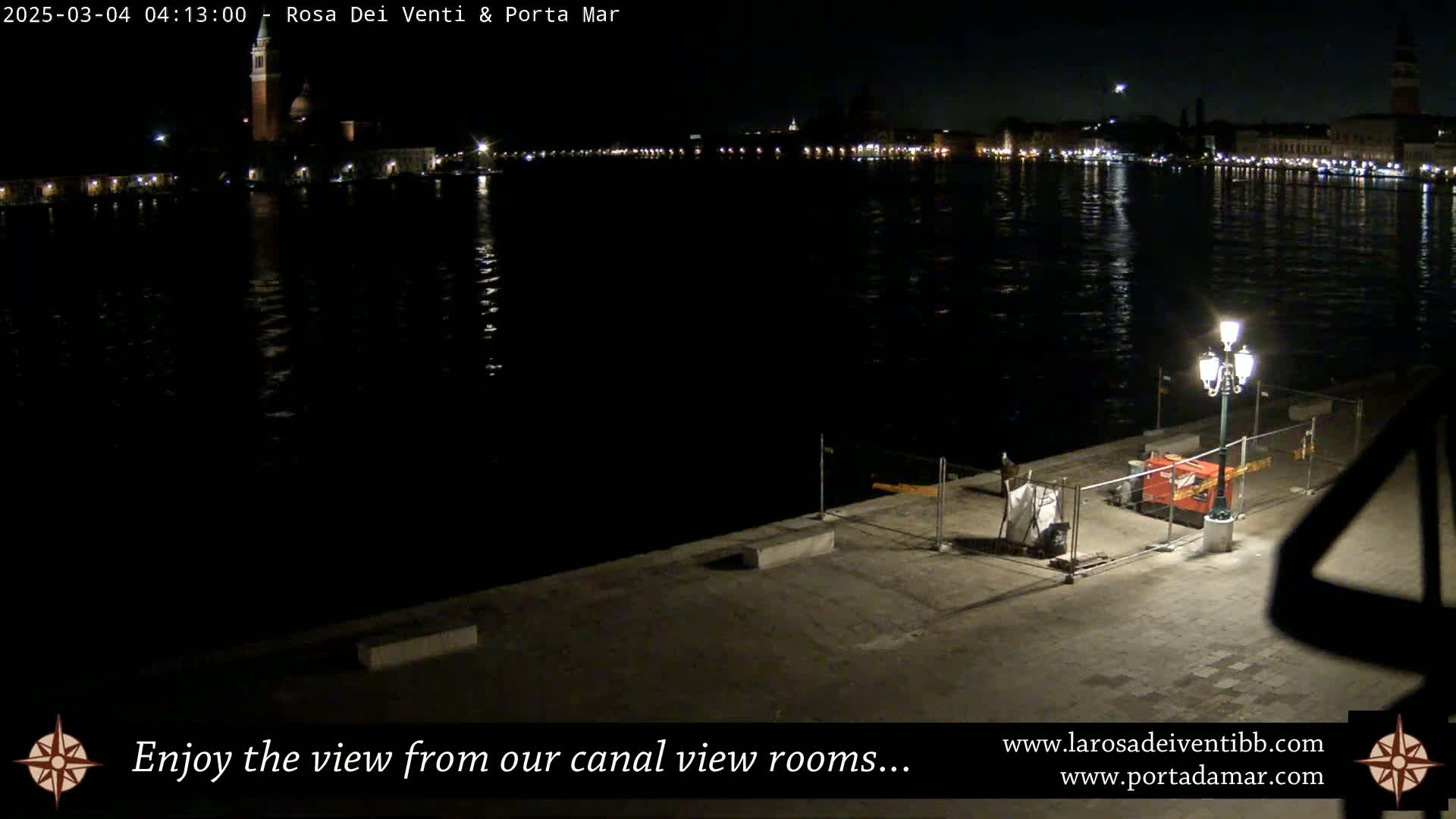 A nighttime view of a canal, reflecting the lights of buildings on the opposite shore, with a lamppost and construction barriers in the foreground.