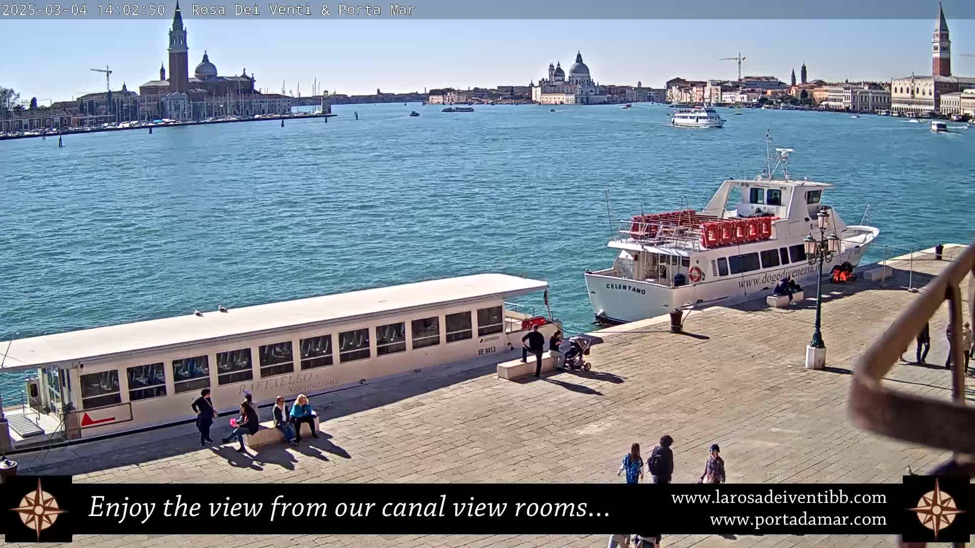 Two passenger boats are docked at a quay, with a view of a calm body of water and a distant cityscape under a sunny sky.