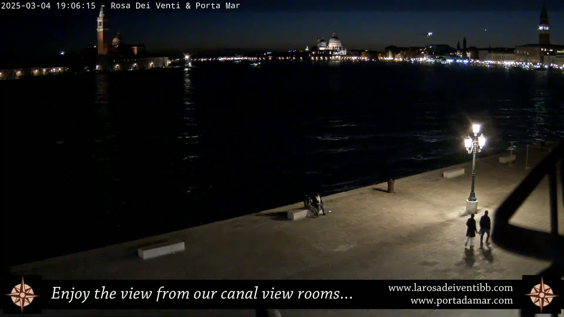 A nighttime view of a calm canal with a city skyline in the background, illuminated by a few lights on the waterfront and a lone lamppost.