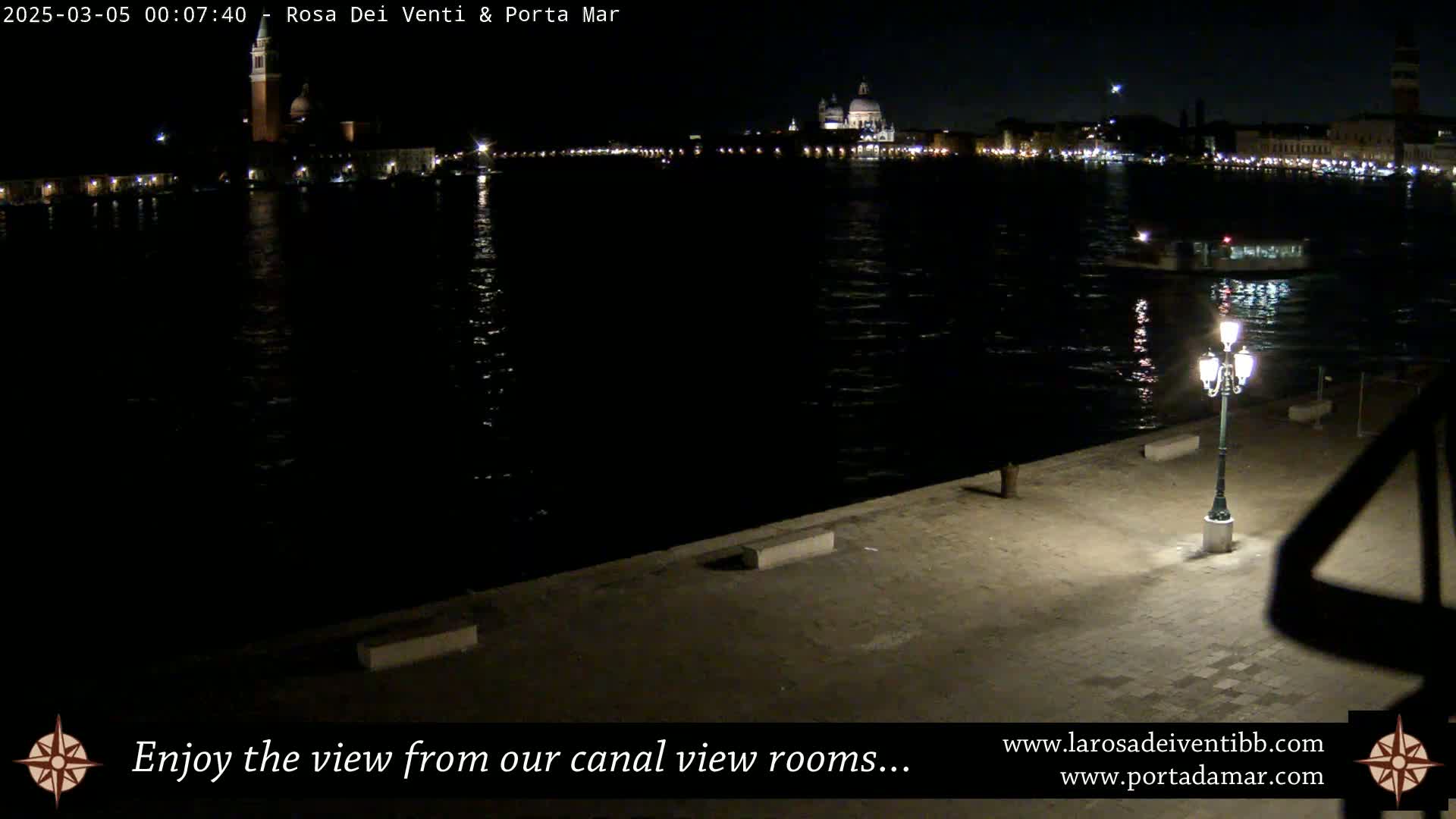 A nighttime view of a calm canal, illuminated by lights on buildings across the water, with a single lamppost on a paved waterfront in the foreground.