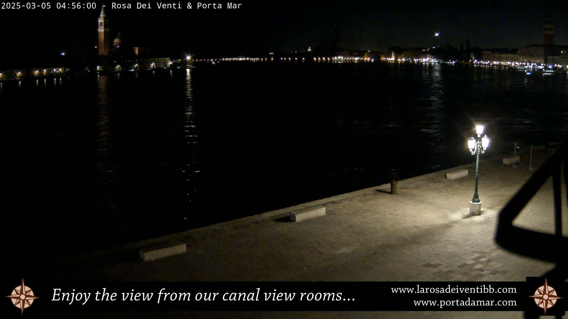 A nighttime view of a calm canal reflecting the lights of a city across the water, with a single lit lamppost on a paved area in the foreground.