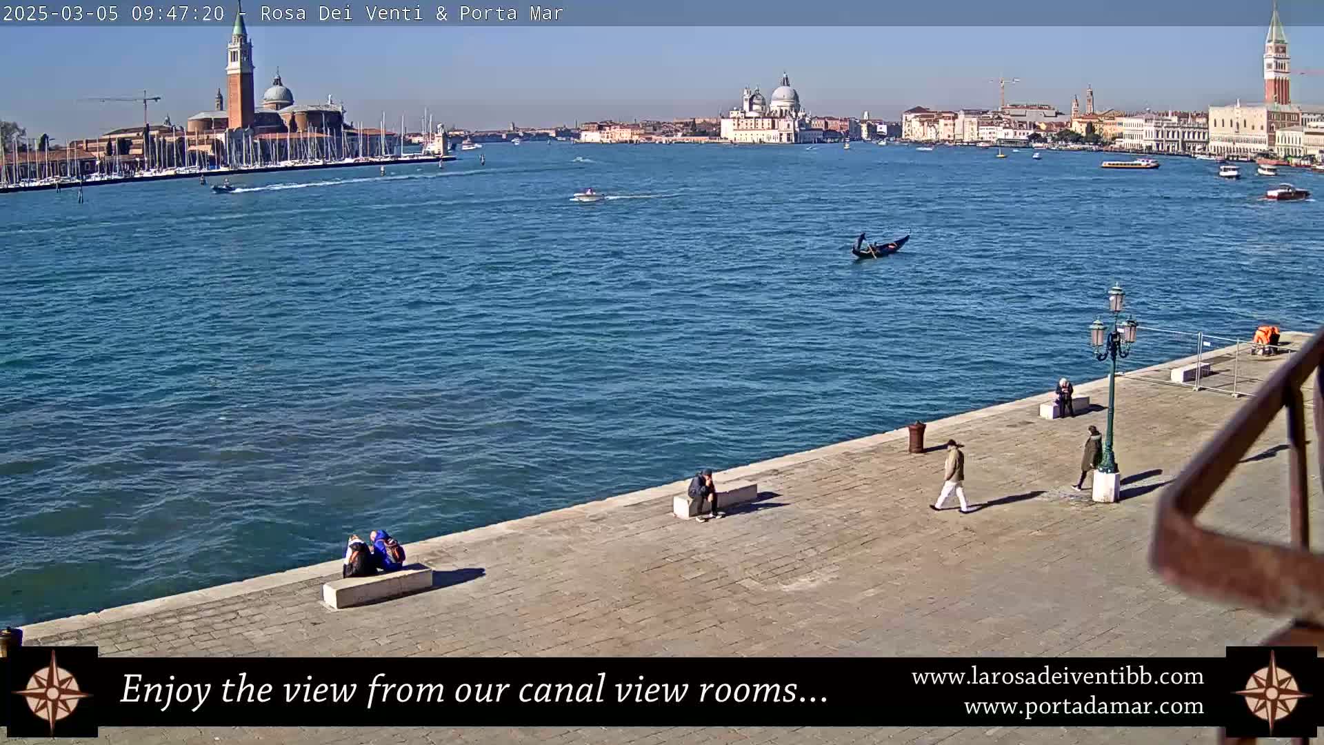 A sunny day in Venice shows a wide expanse of blue water with boats and gondolas, a cityscape across the water featuring prominent buildings, and a few people walking along a stone embankment.