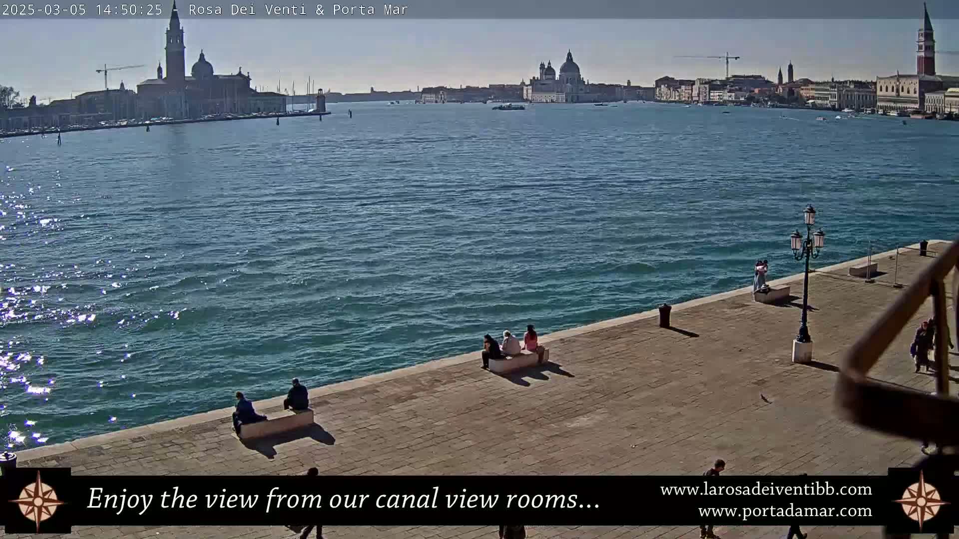 A sunny day in Venice shows a canal view with several people sitting on a stone embankment overlooking the water and distant buildings.