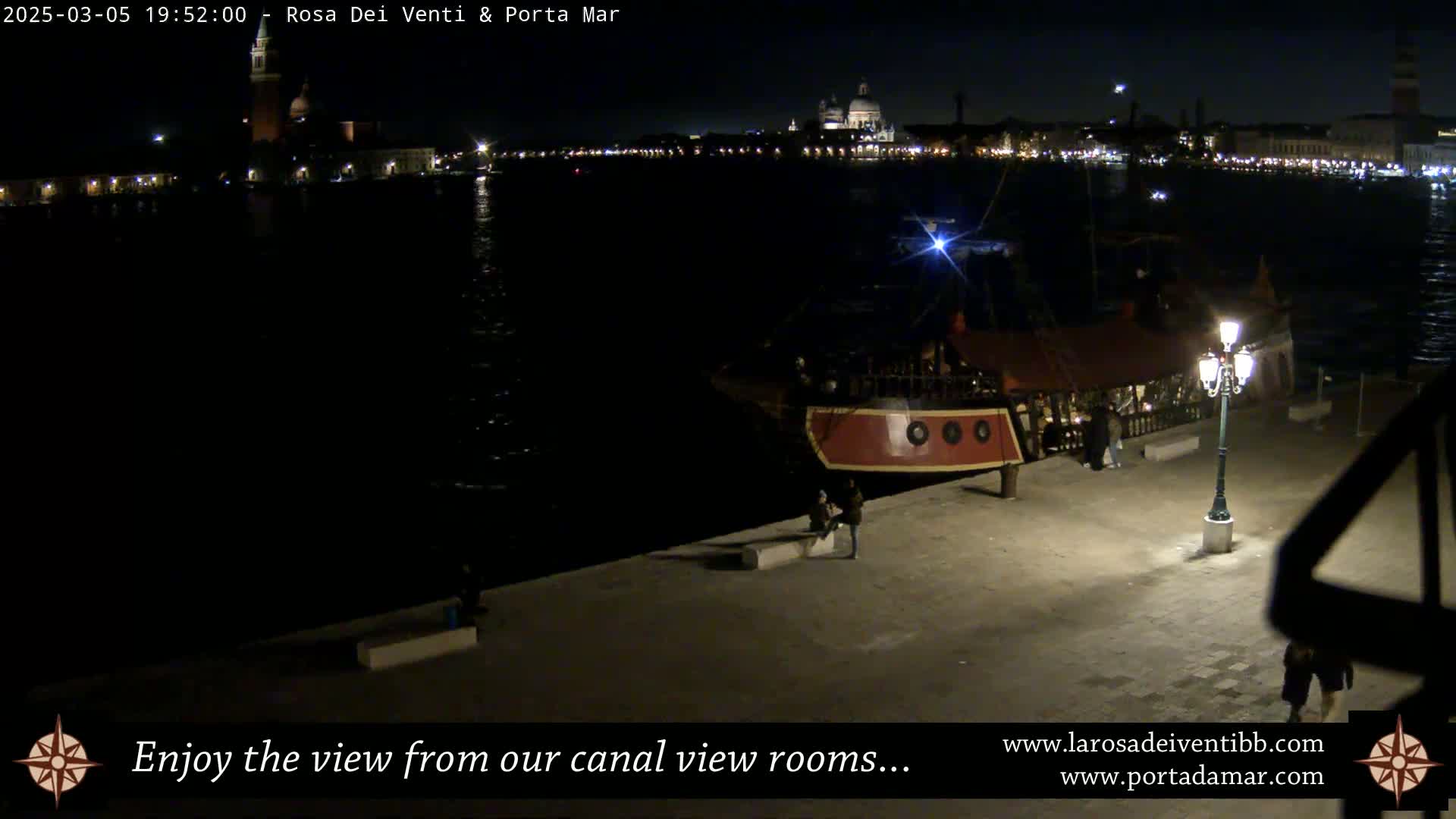 A nighttime view of a canal in Venice, Italy, showing a docked boat, a waterfront walkway, and city lights across the water.