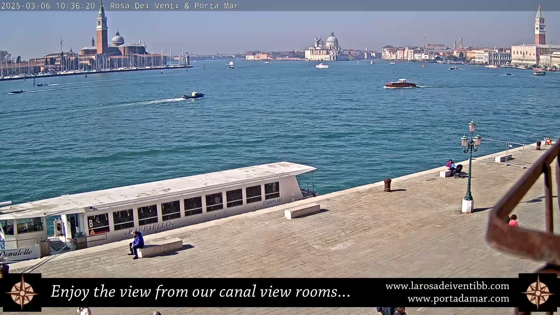A sunny day in Venice shows a canal with boats, a docked passenger vessel, and a waterfront paved area with people; in the distance, are Venetian buildings.