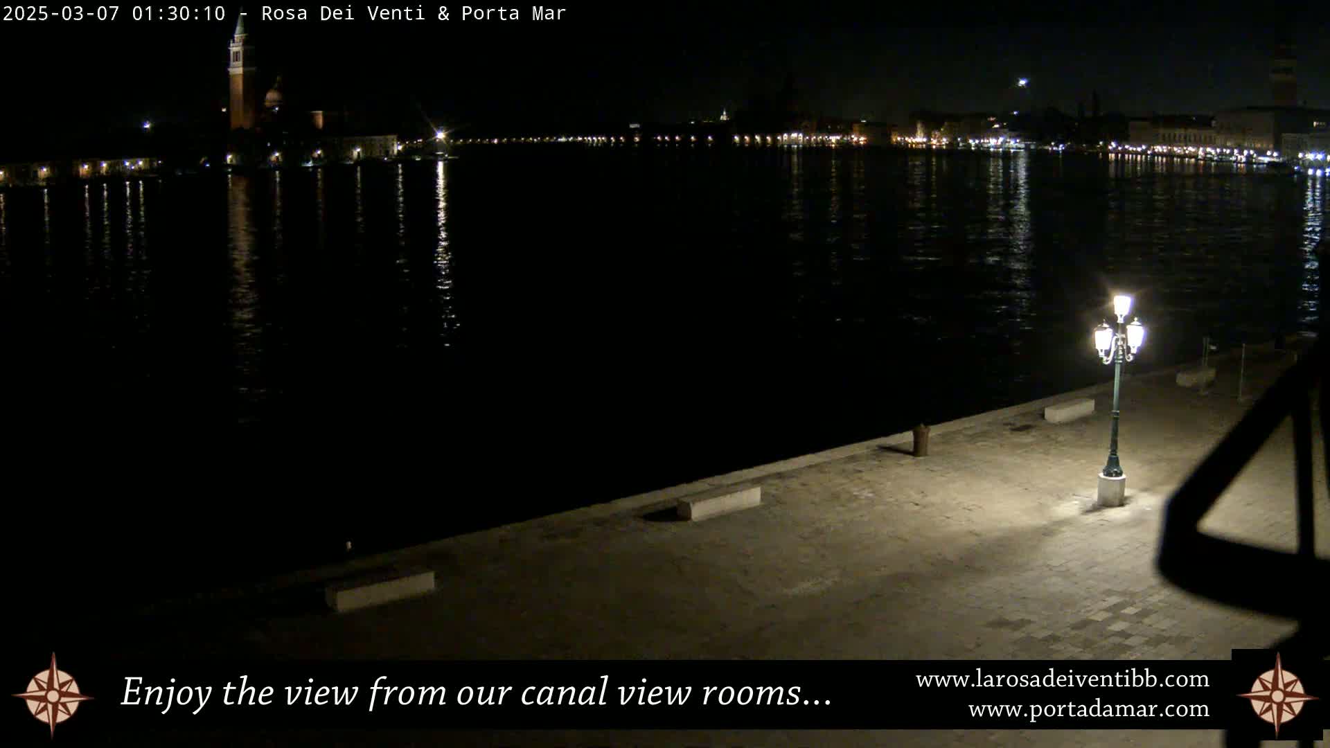 A nighttime view across a calm canal shows the lights of a city across the water, with a single lit lamppost on a paved waterfront in the foreground.