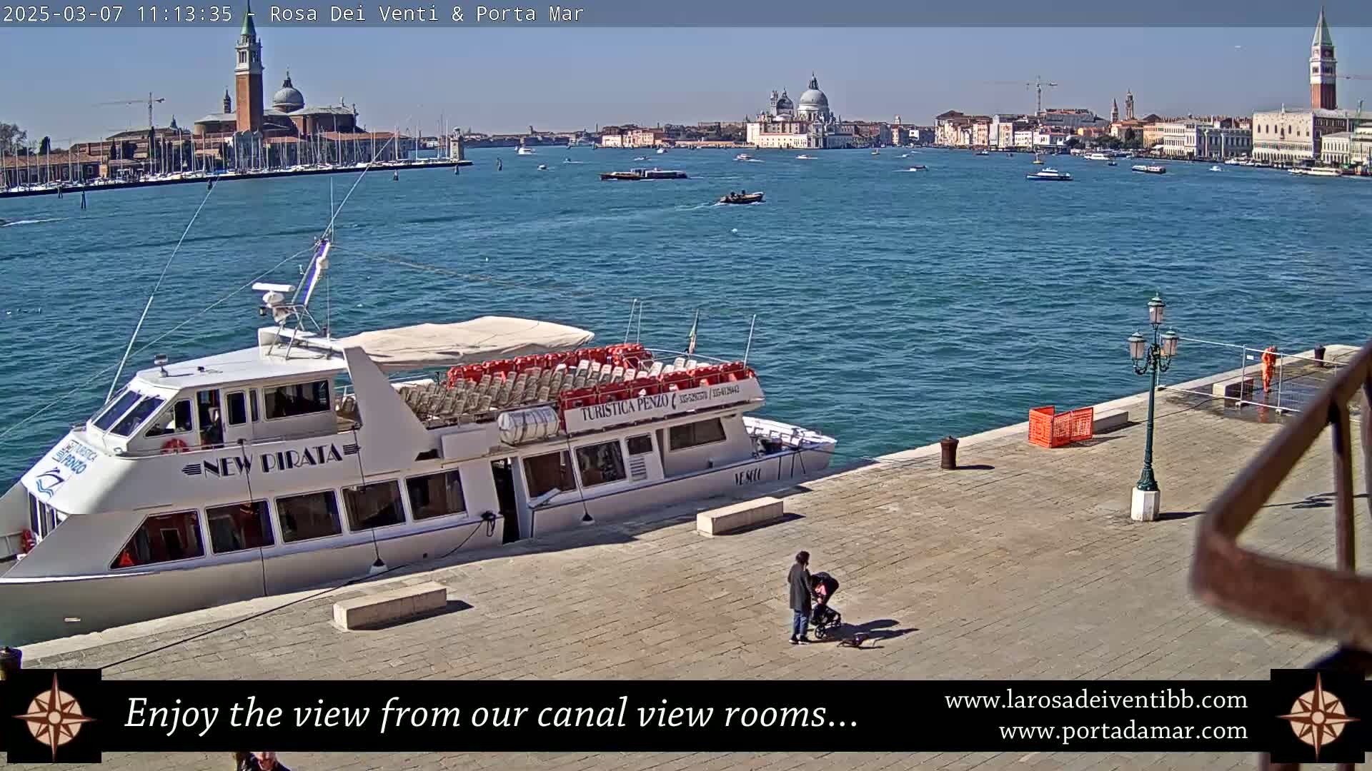 A white passenger boat is docked at a waterfront paved area overlooking a calm blue body of water with distant Venetian buildings under a sunny sky.