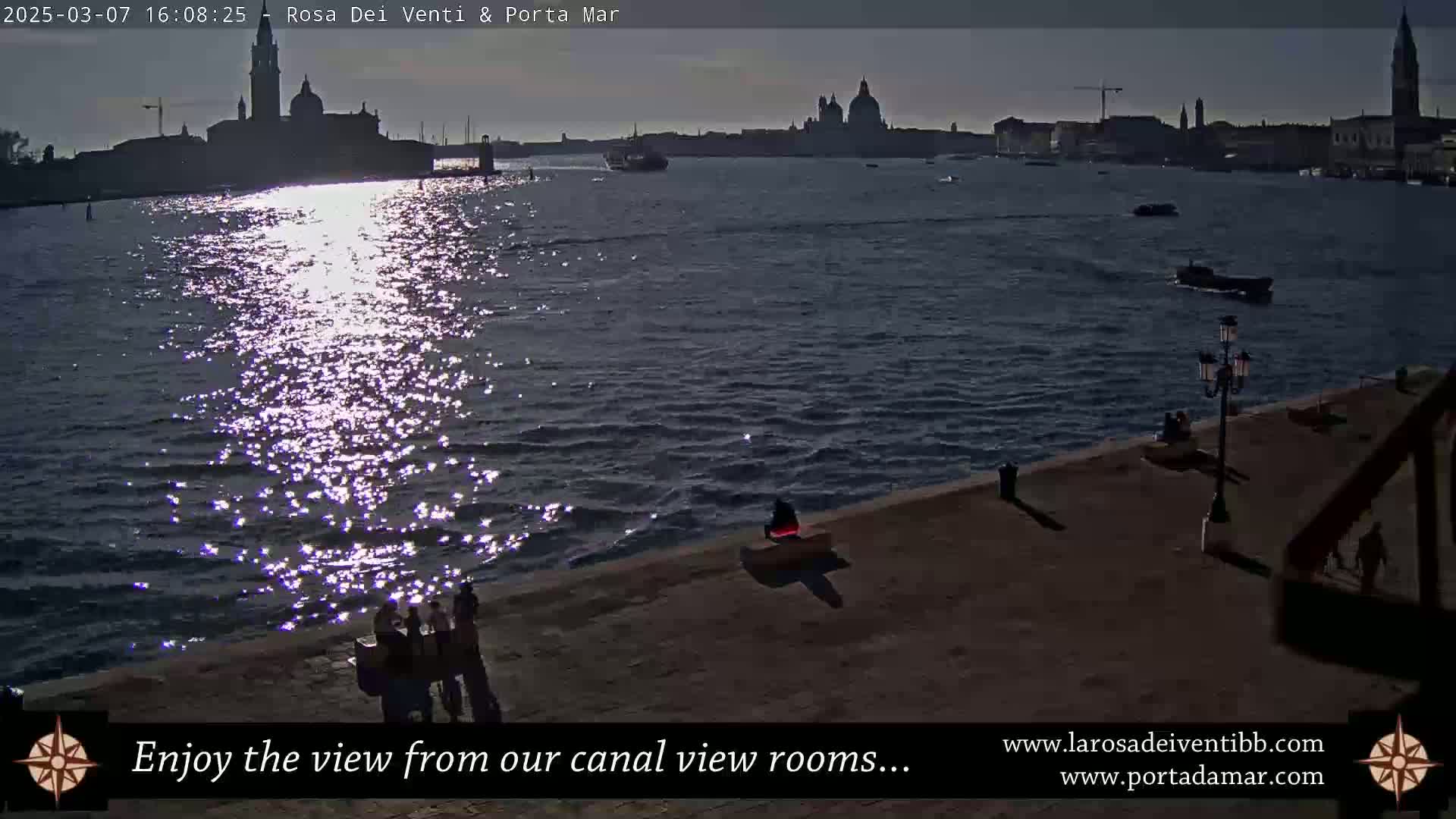 A sparkling canal reflects the setting sun, illuminating the silhouetted cityscape across the water, where a few people sit along a paved waterfront.