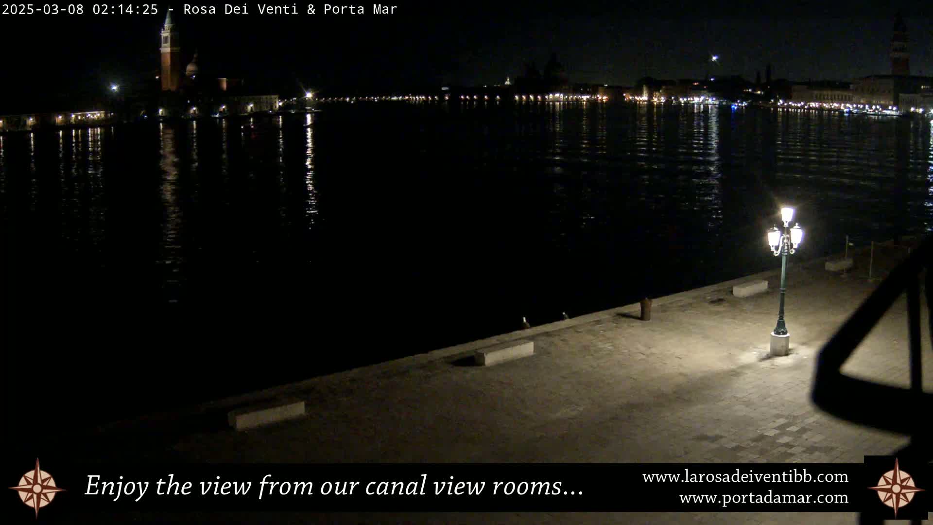 A nighttime view of a calm canal reflecting city lights, with a lit lamppost on a paved waterfront.