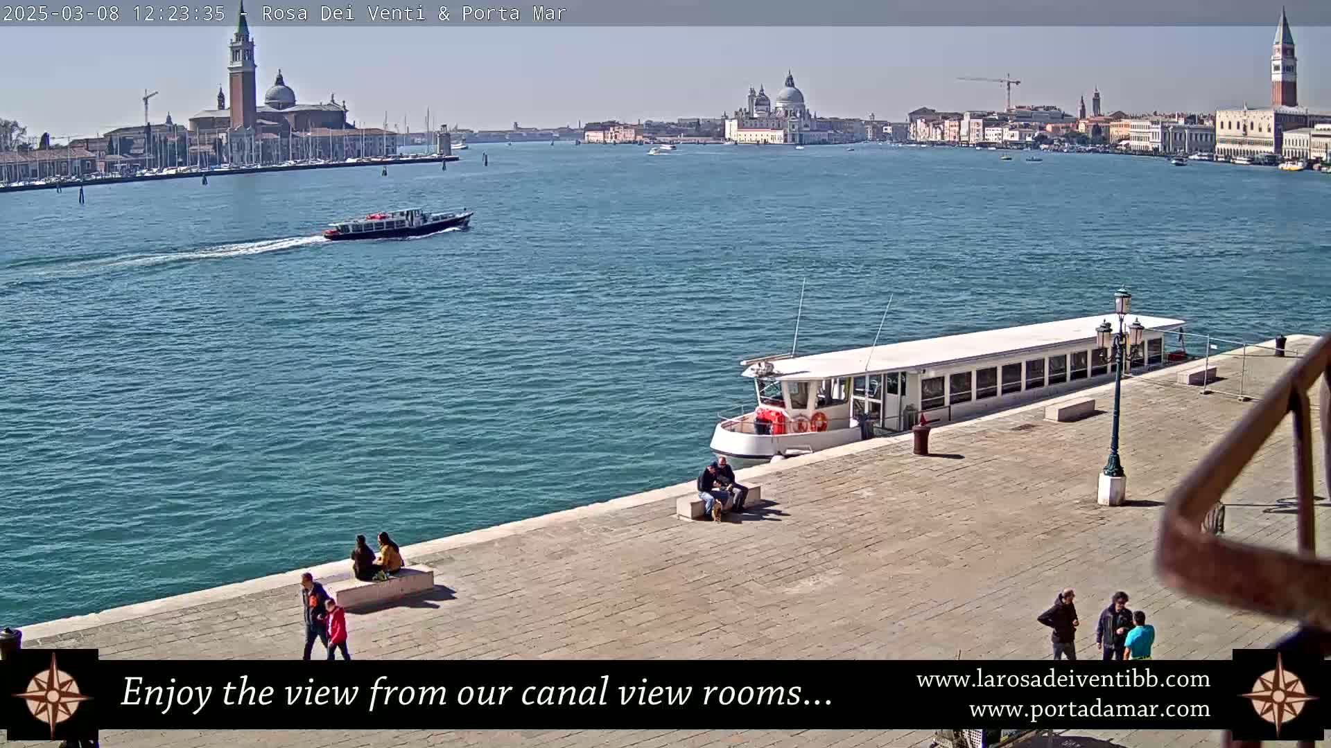 A sunny day in Venice shows a wide canal with boats, several people walking along a paved waterfront area, and a cityscape of buildings and churches in the background.
