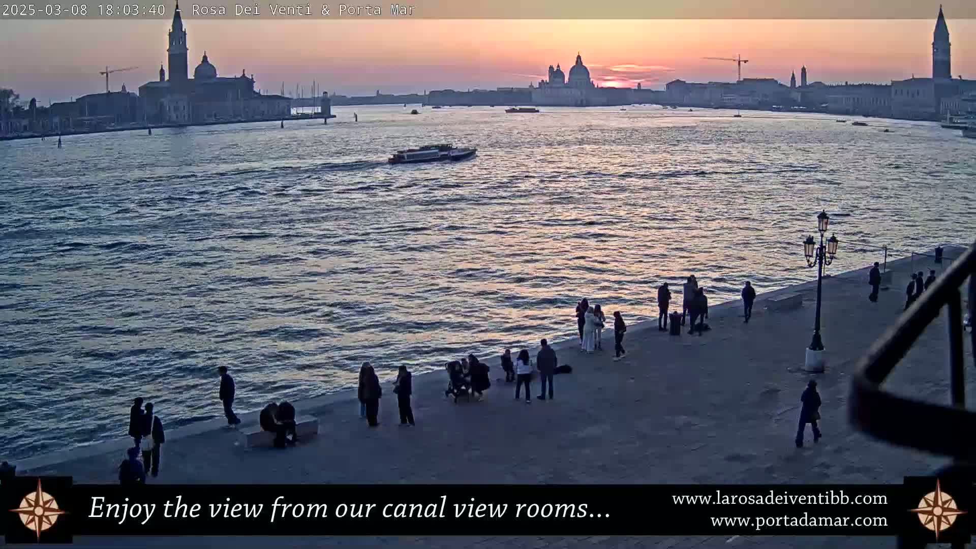 A twilight view of a calm canal with a group of people standing on a waterfront walkway, overlooking a city skyline with several prominent buildings, including a large domed structure.