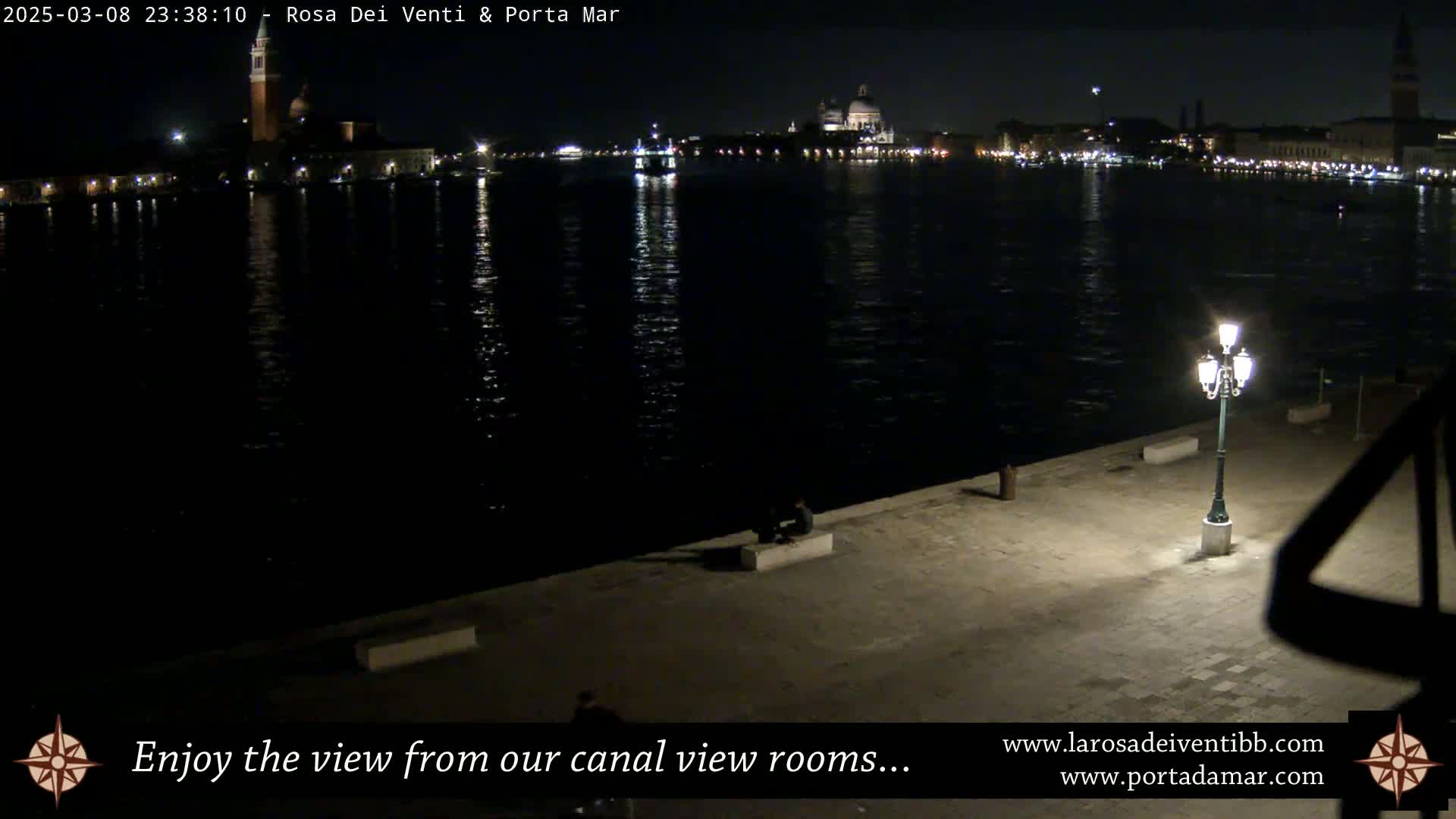 A nighttime view of a calm canal in Venice, showing city lights and buildings across the water, with a few people visible on a nearby embankment.