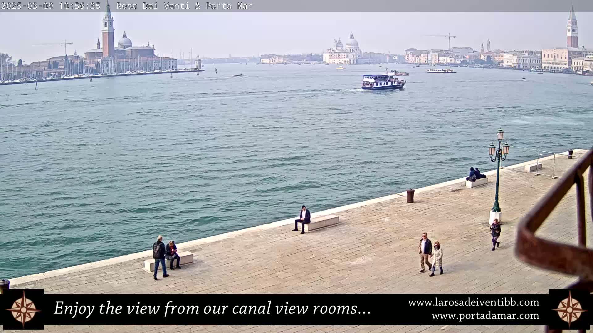 A calm canal scene in Venice shows a waterway with several boats, a cityscape including a large church, and a few people walking and sitting along a paved waterfront on a hazy day.