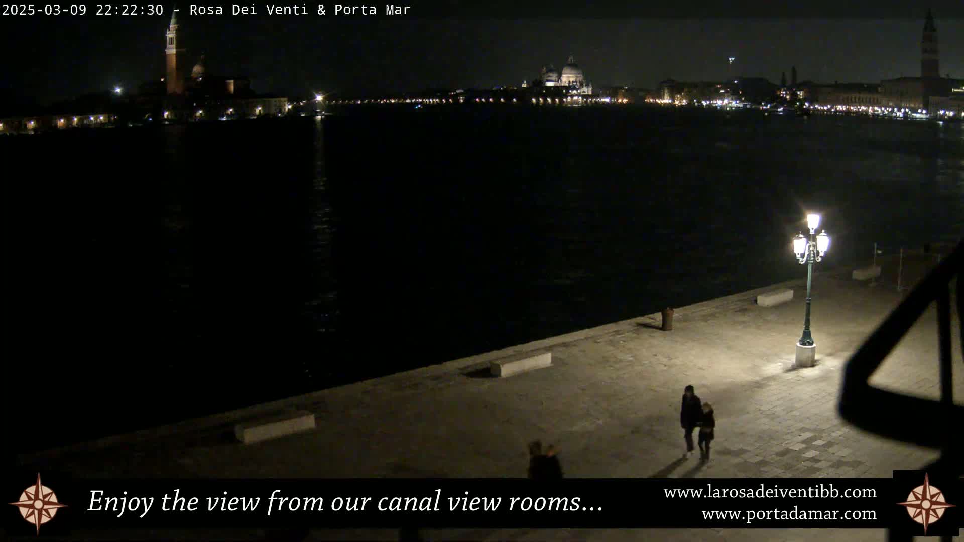 A nighttime view of a calm canal in Venice, showing illuminated buildings across the water and two people walking on a paved area in the foreground, under a lit lamppost.