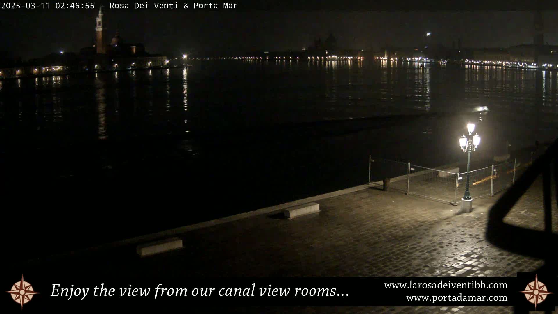 A nighttime view of a calm body of water with lights illuminating the far shore and a single lamppost on a cobblestone walkway in the foreground.