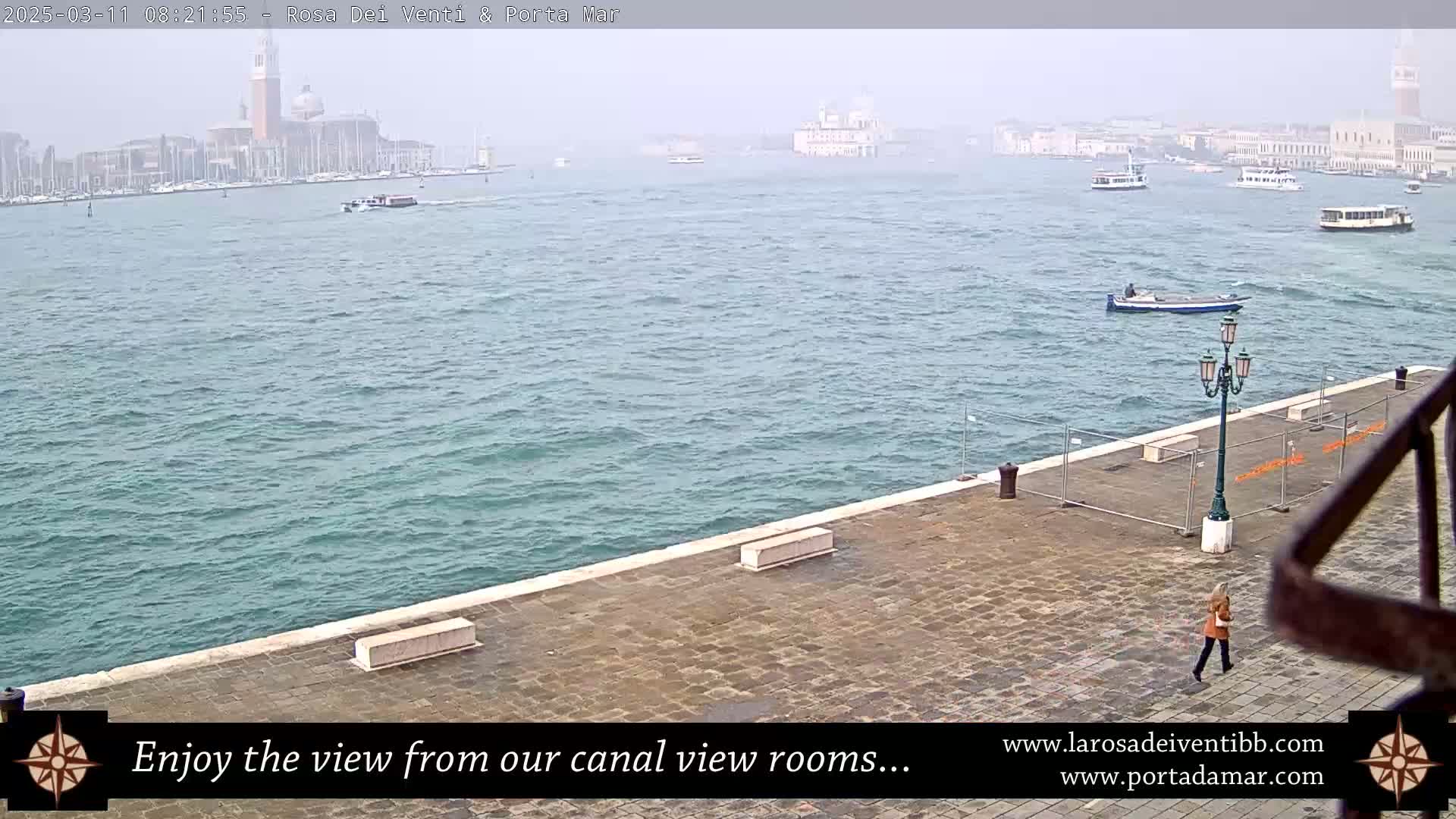 A hazy view of a waterway shows boats and buildings in the distance, with a stone walkway and a person walking in the foreground.
