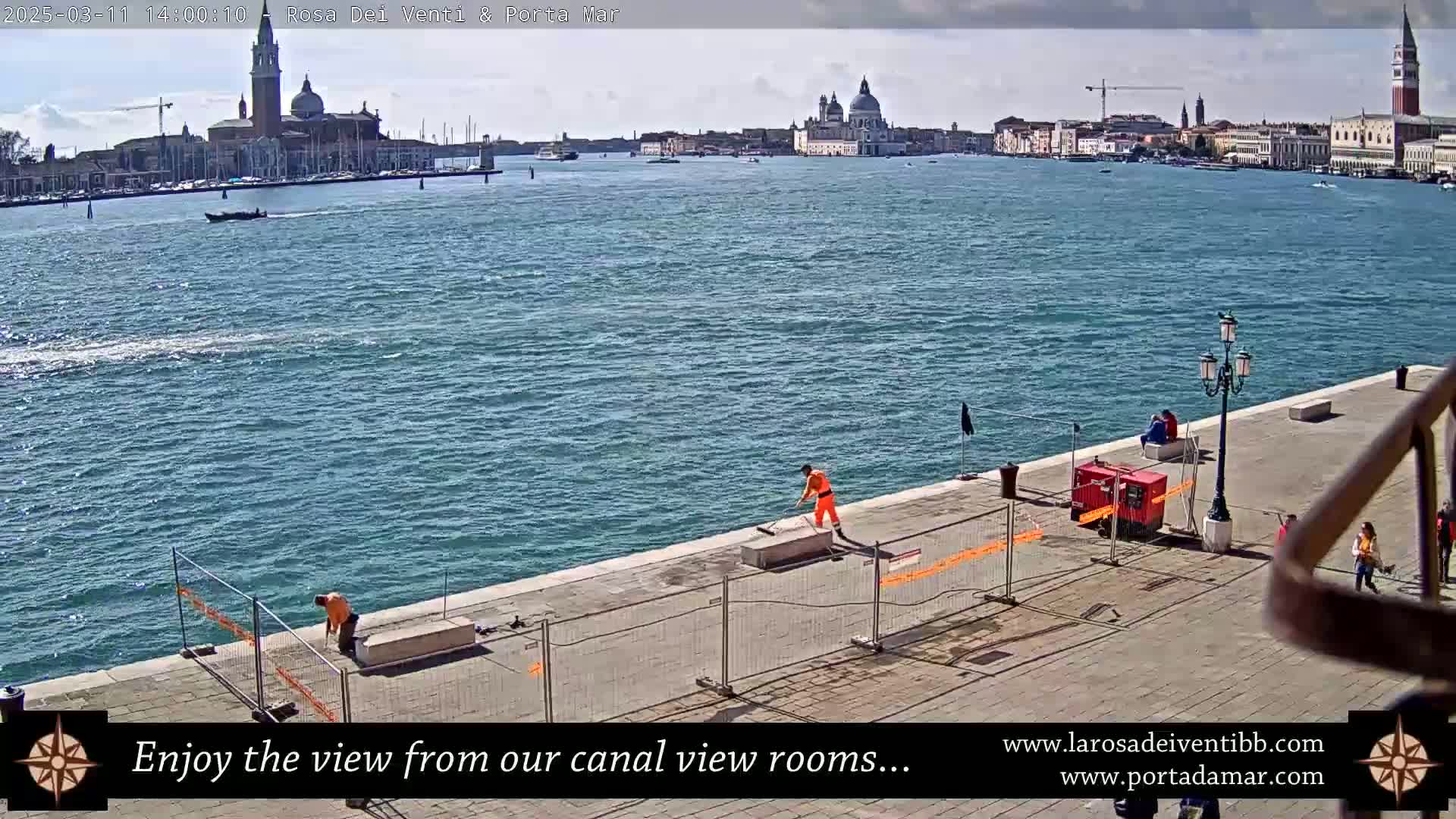 A sunny day shows a waterway with a city skyline in the distance, workers cleaning a paved waterfront area, and people sitting and walking nearby.
