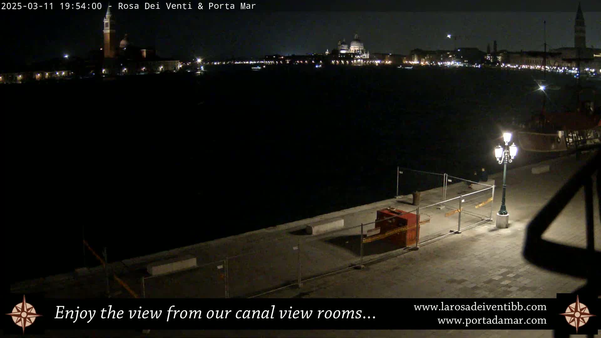 A nighttime view of a canal, showing a cityscape with illuminated buildings across the water, and a paved waterfront area with construction fencing and a lamppost in the foreground.