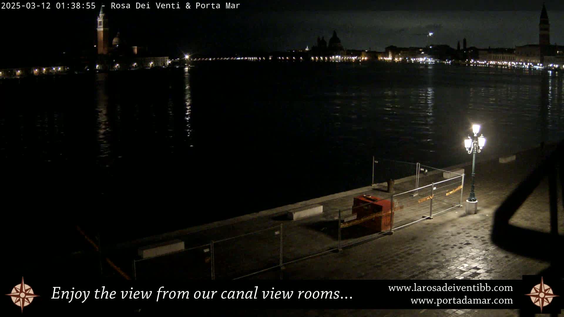 A nighttime view across a calm canal shows a city skyline with illuminated buildings and a single lit lamppost on a paved waterfront area partially blocked by a temporary fence.