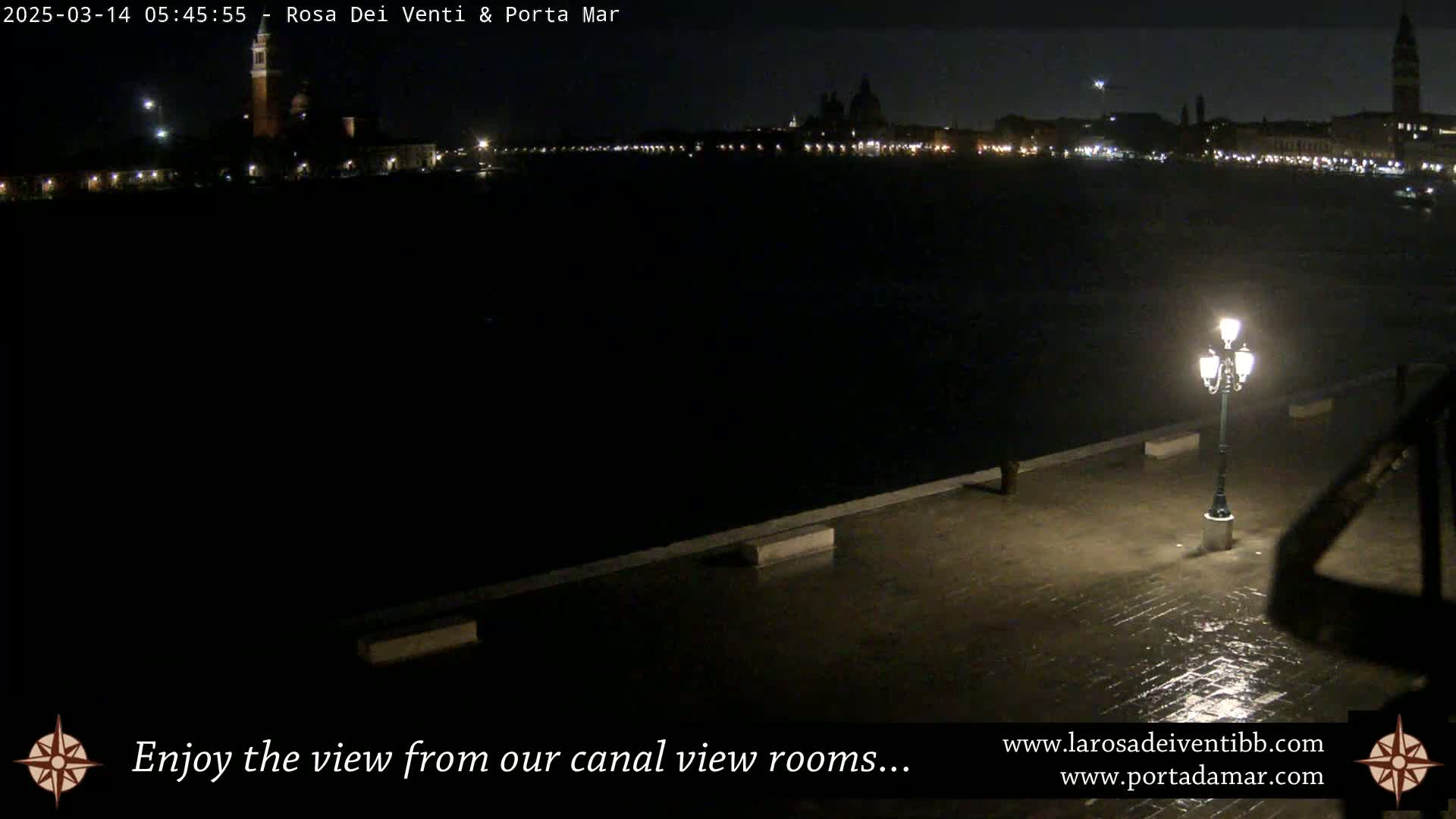 A nighttime view of a canal, showing a cityscape with lights across the water, and a wet, paved area with a lit lamppost in the foreground.