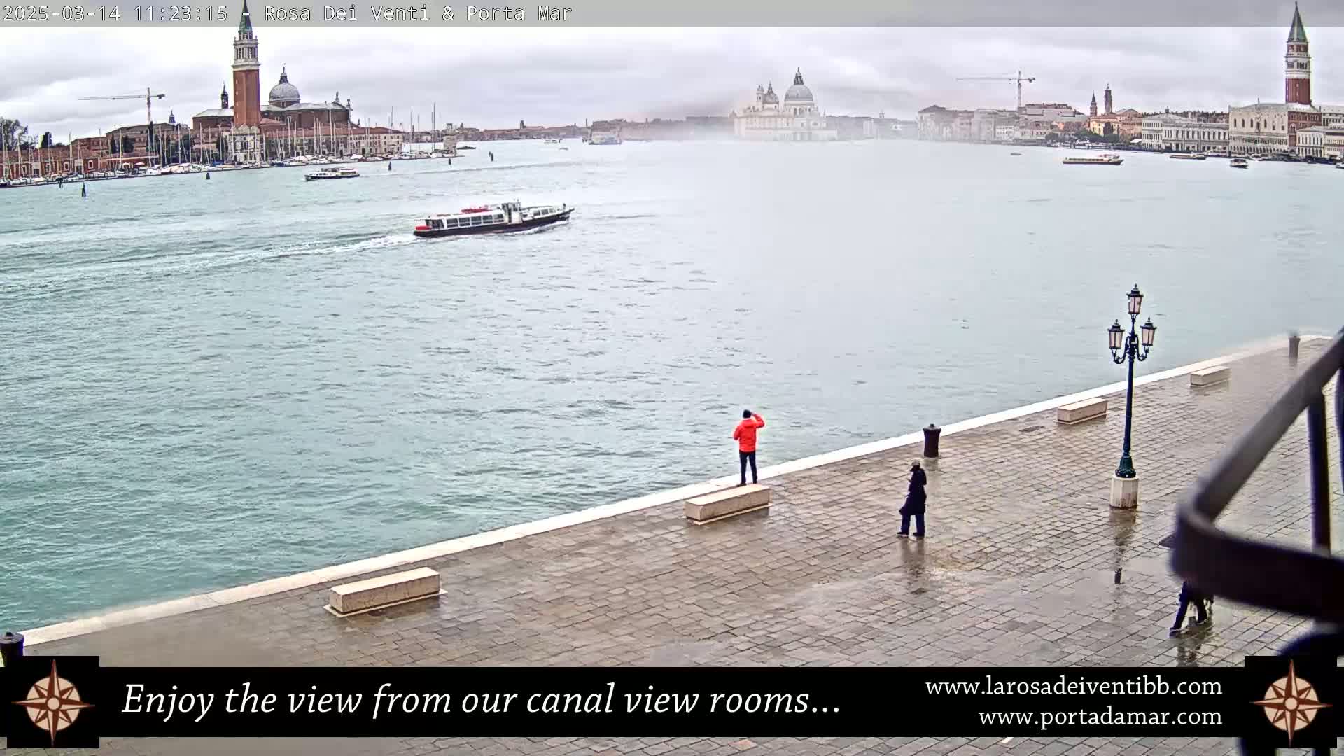 A wide shot of a waterway shows a city in the distance under an overcast sky, with a few people standing on a paved waterfront area in the foreground.
