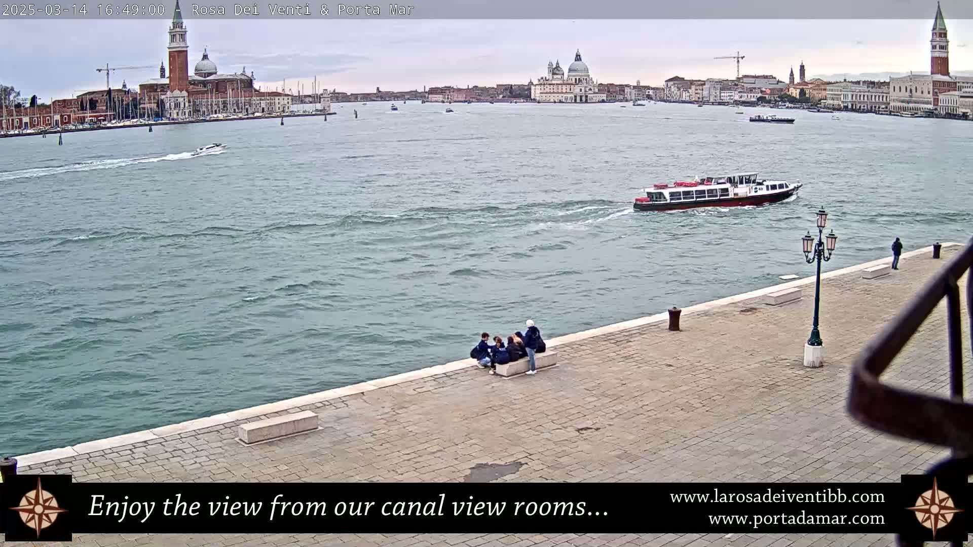 A waterfront view of a canal in Venice, Italy, on an overcast day shows several people sitting on a stone embankment with a boat in the water and a city skyline in the distance.