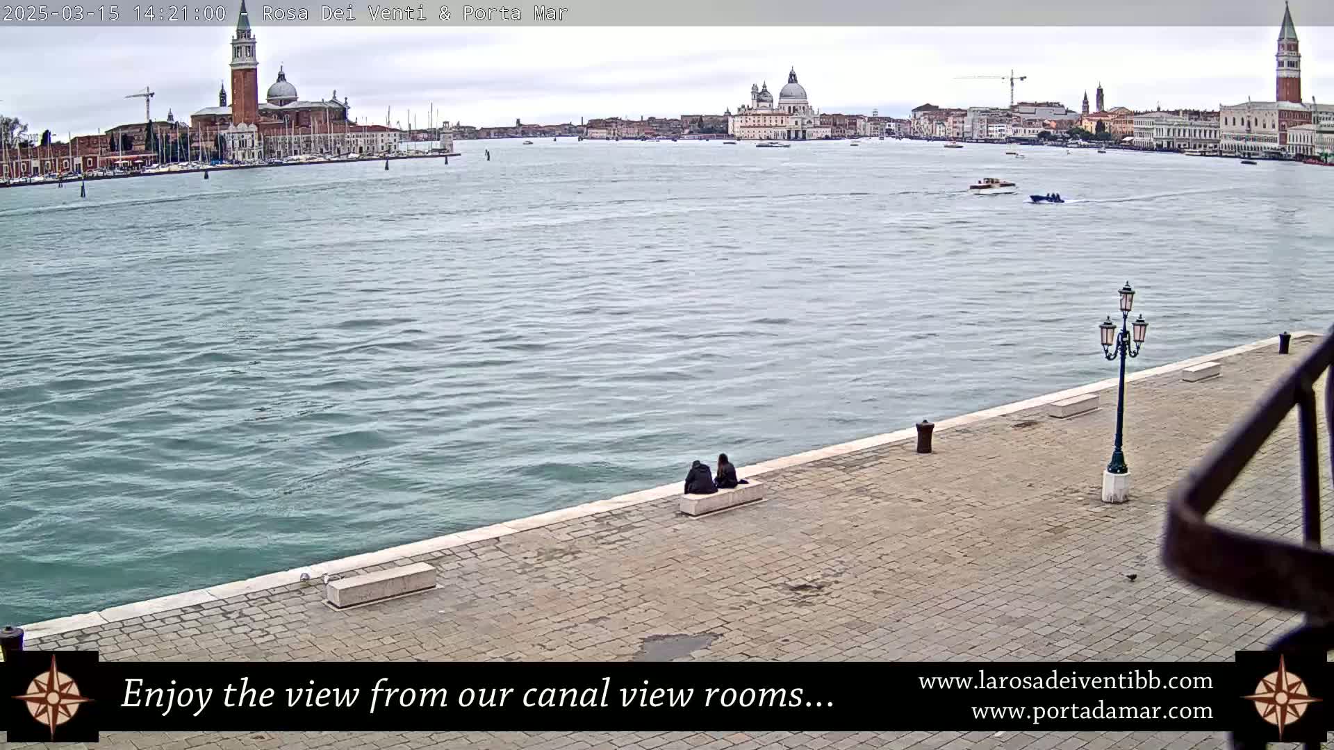 A wide shot of a calm waterway shows a city skyline across the water and two people sitting on a low wall along the shore on an overcast day.