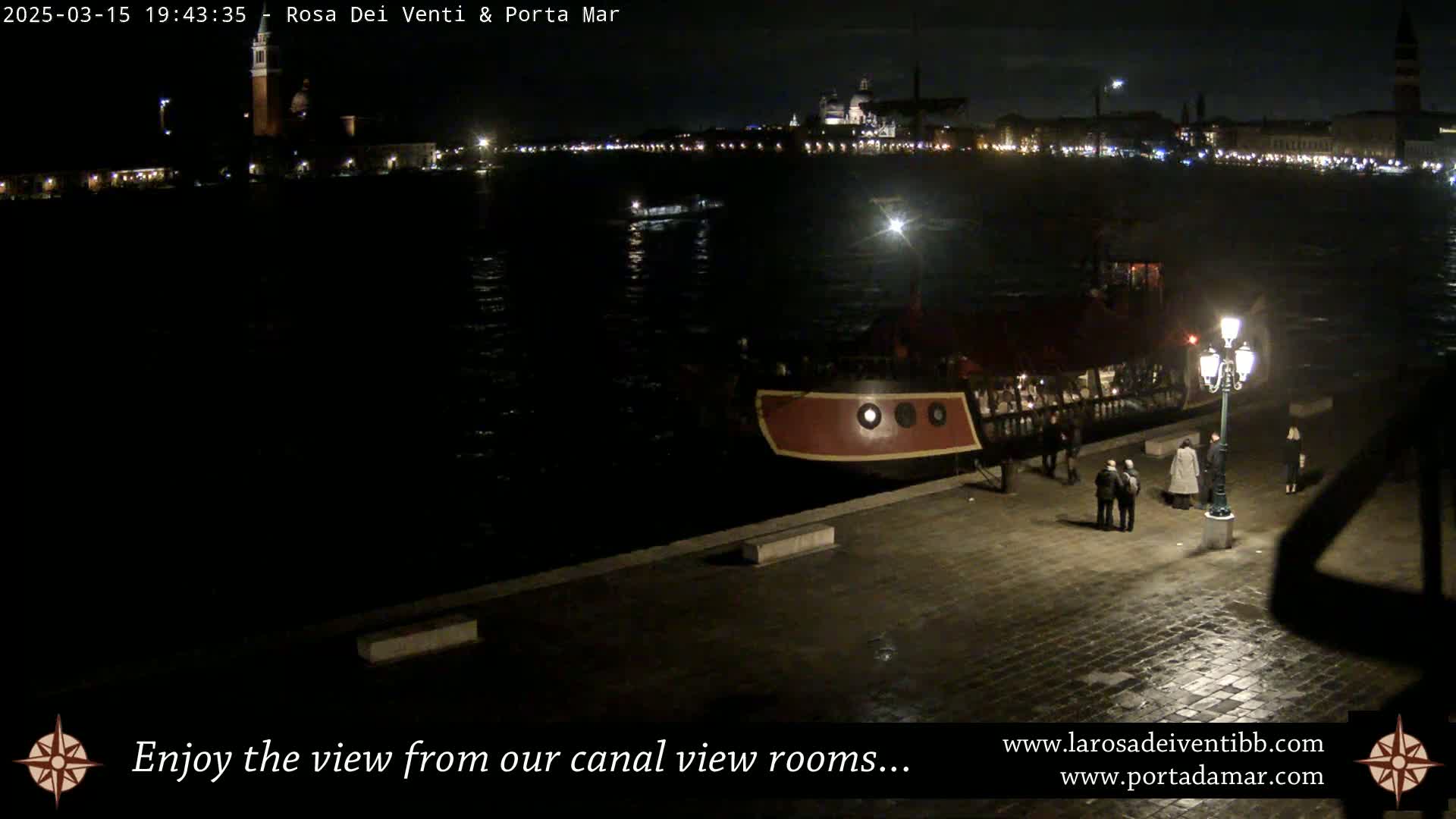 A nighttime view of a canal shows a docked red boat with people standing nearby,  across from a city skyline illuminated by lights.