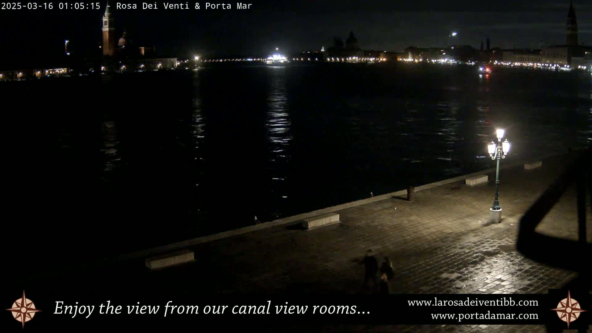 A nighttime view of a calm canal reflecting the lights of a city across the water, with a stone walkway and a lit lamppost in the foreground.