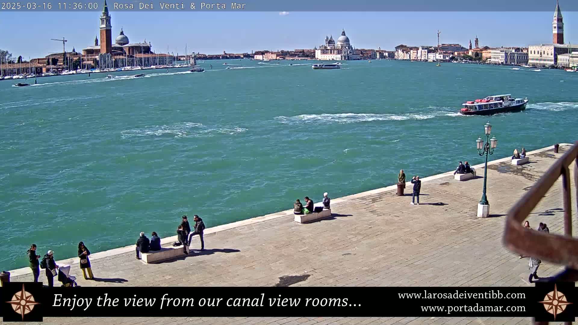 A sunny day in Venice shows a waterfront paved area with several people sitting and standing, overlooking a waterway with boats and a distant cityscape featuring prominent buildings.