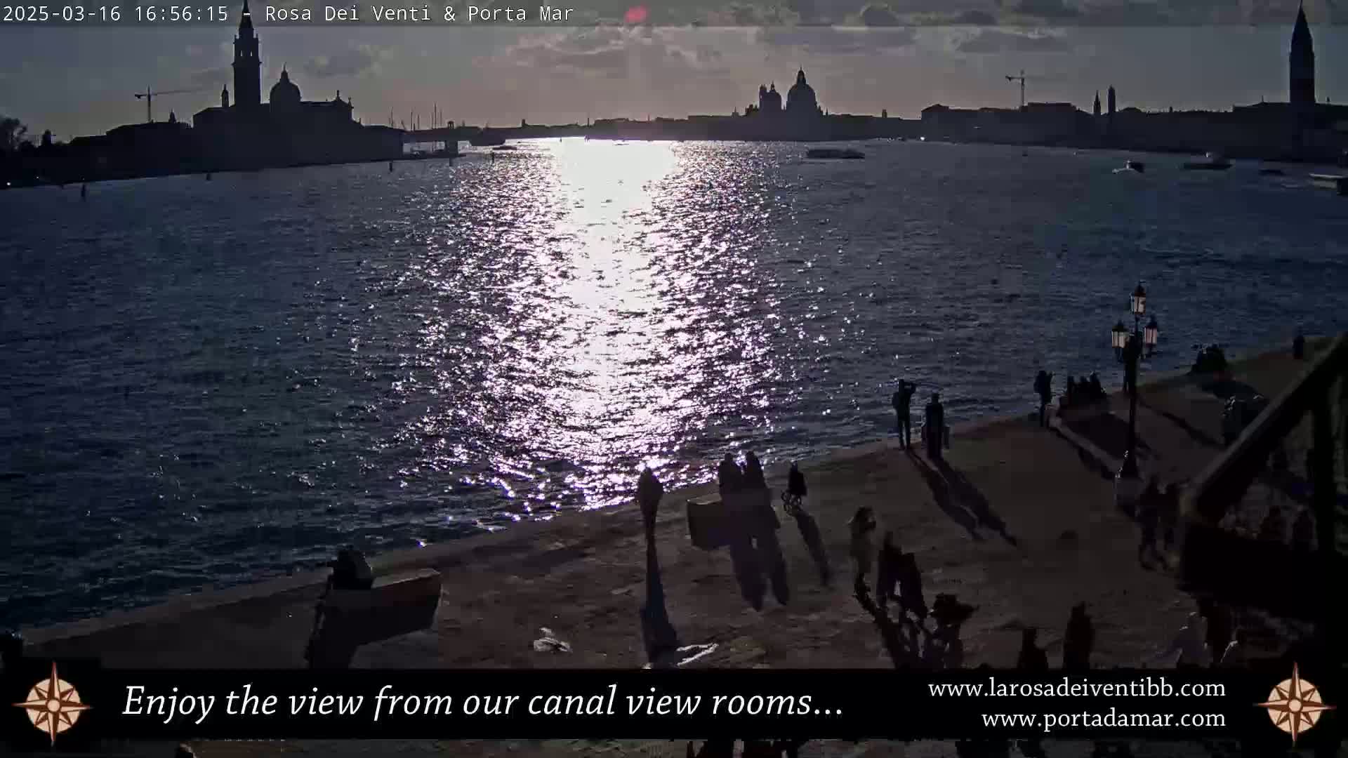 A silhouetted cityscape is visible across a shimmering waterway at sunset, with several people standing along a waterfront walkway casting long shadows.