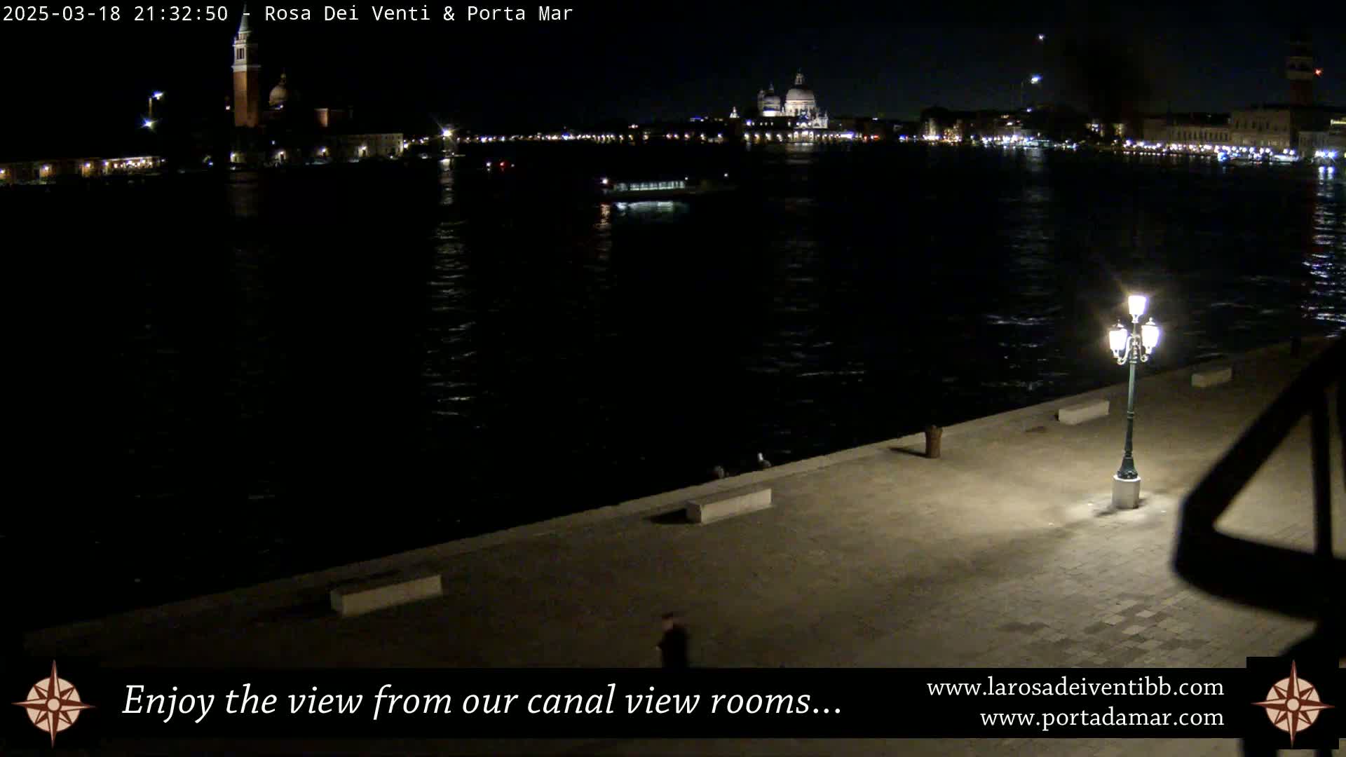 A nighttime view of a canal, with city lights and a building with a dome in the distance, seen from a paved waterfront area with a single lit lamppost.