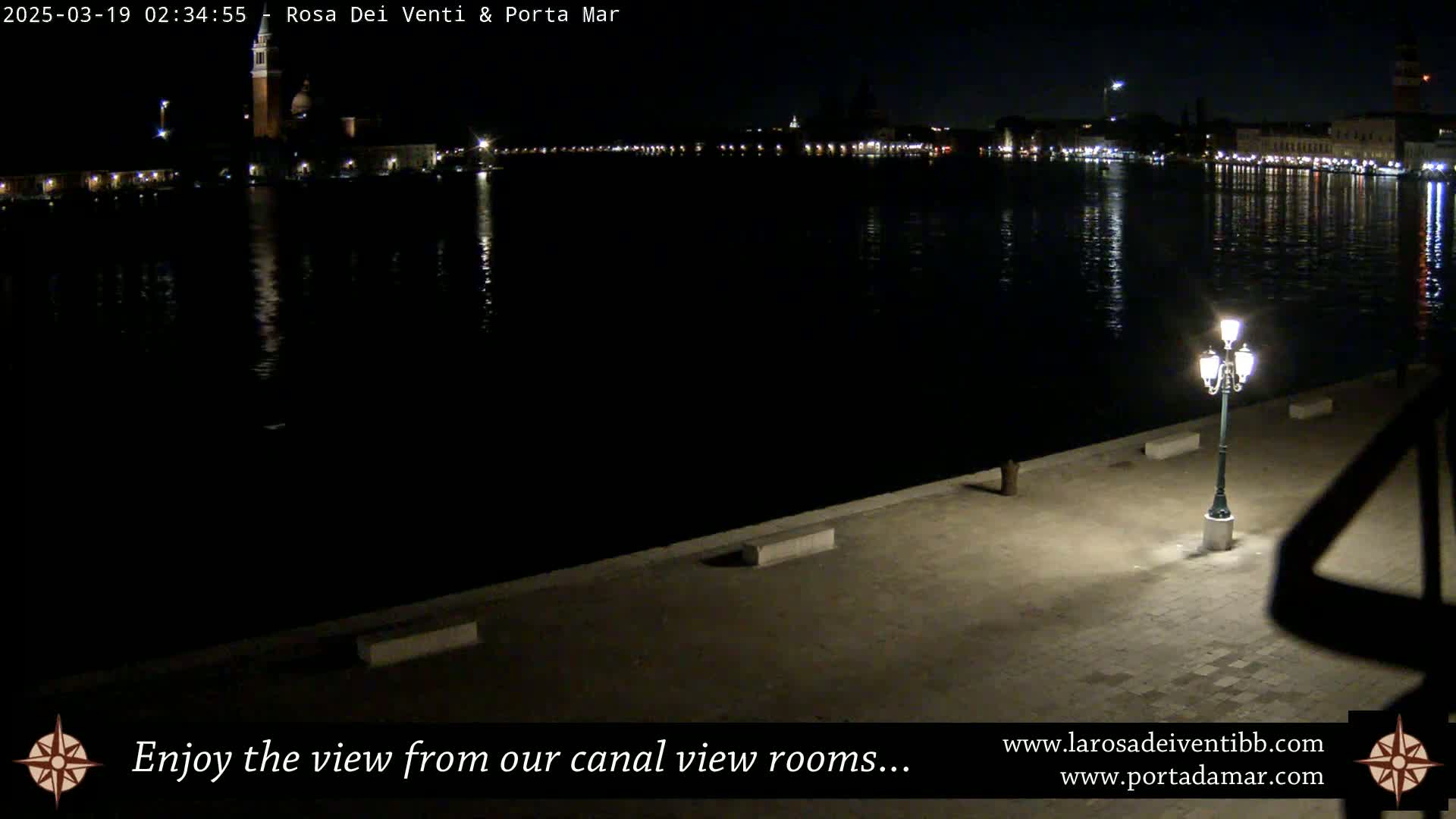 A nighttime view of a calm canal reflecting illuminated buildings across the water, with a single lamppost on a paved area in the foreground.